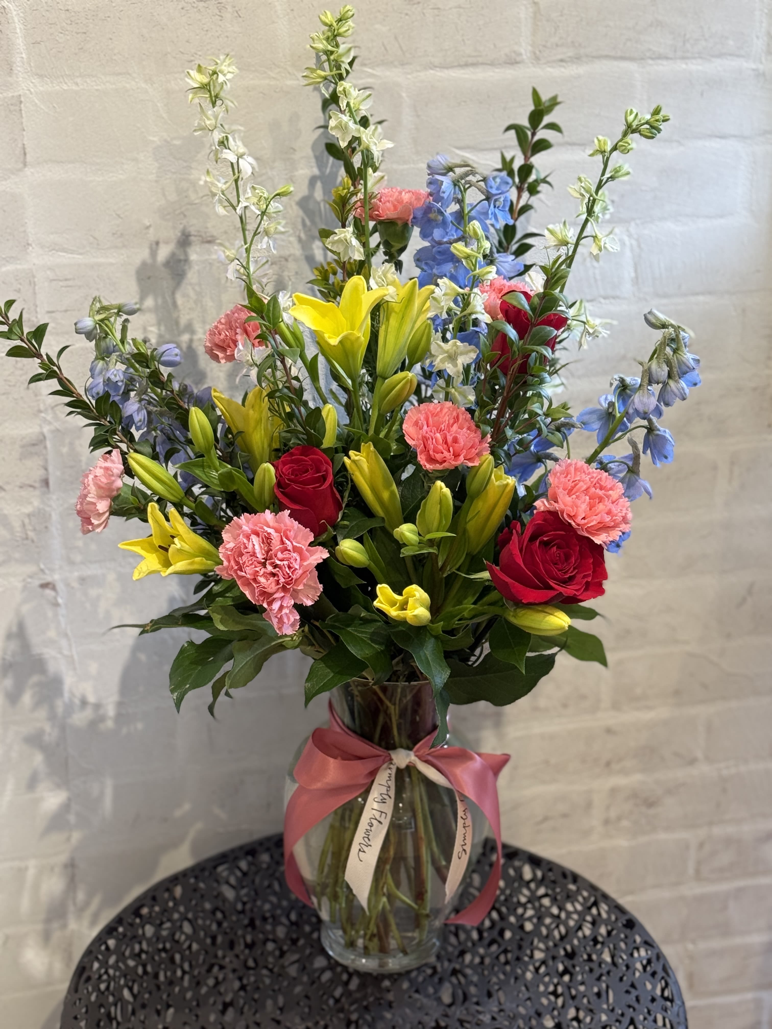 New! Petal Perfection - A tall, front facing linear arrangement of mixed colors. Lilies, roses, delphinium and more. Measuring approximately 30” tall. 