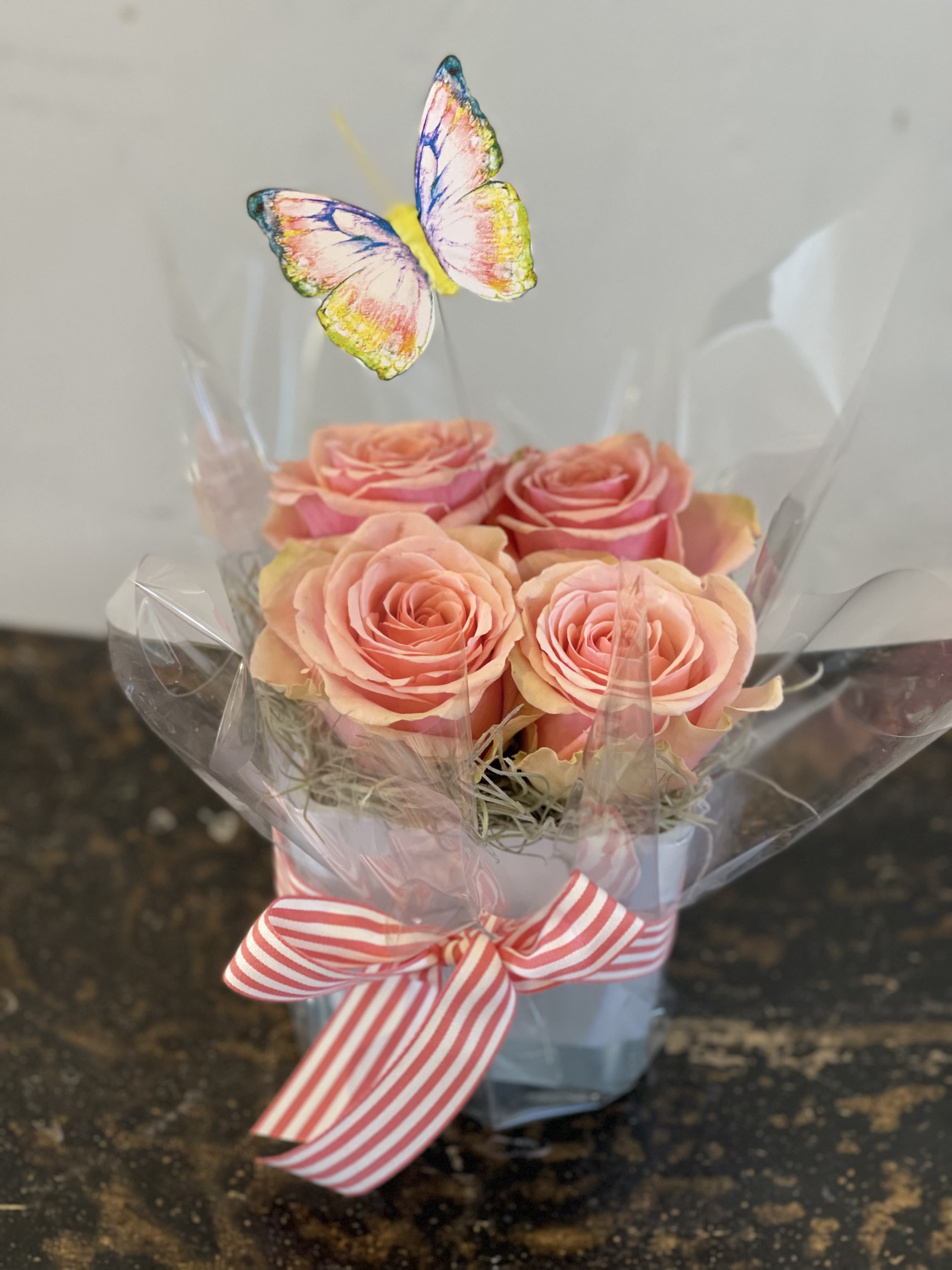 Butterfly Kisses -  A square vessel filled with 4 pave roses. Finished off with a butterfly, moss and cello wrap. All tied up with a bow. Rose color may vary. Want a specific color? Let us know in the notes! 