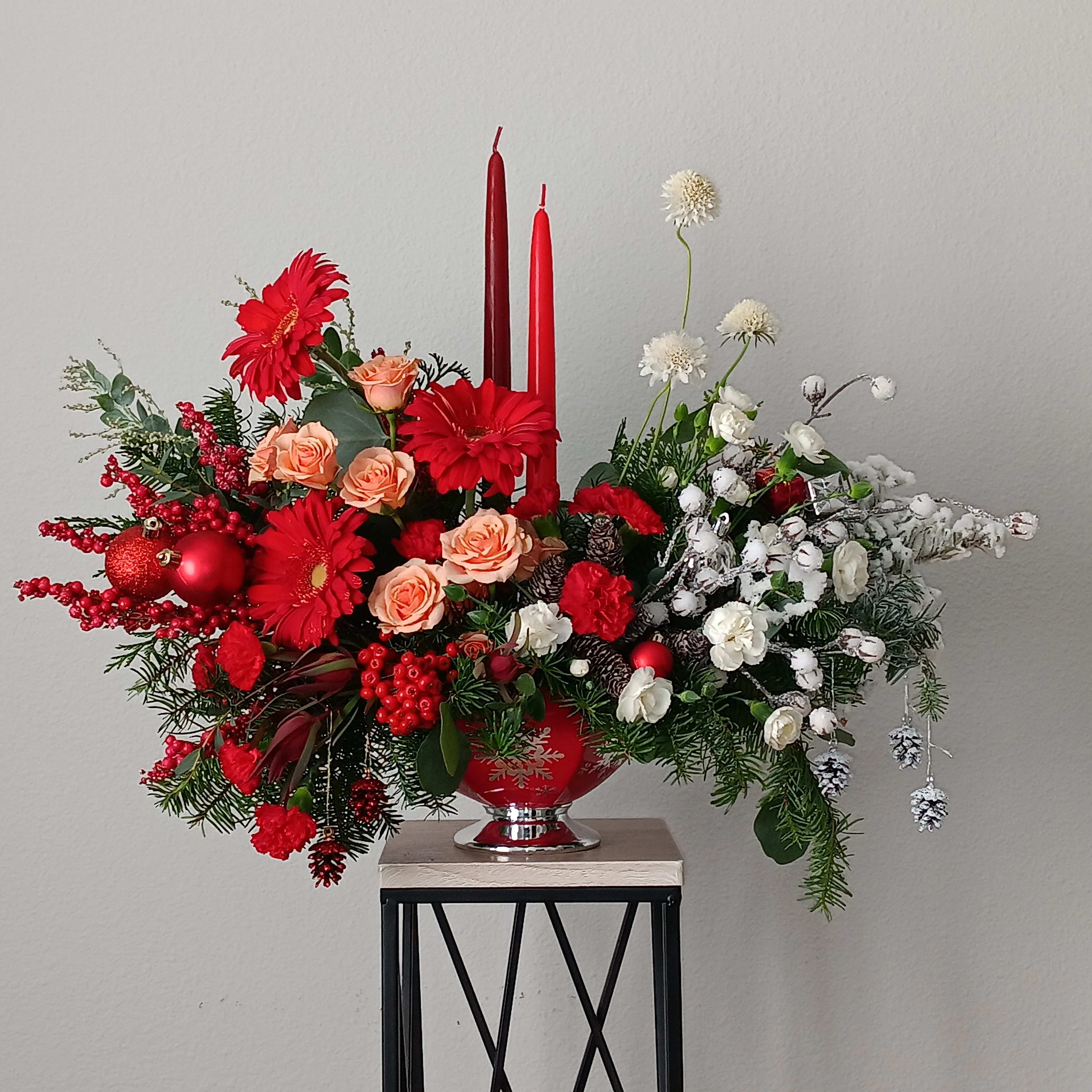Merry Magnificence - DFS - A magnificent arrangement for any holiday event. As a beautiful Christmas centerpiece or mantle decoration, this unique arrangement mixes  red gerbera, red mini carnations, white   scabiosa, white spray roses, with festive greens around the base of a glowing pillar candle.  Orientation: All-Around  SUBSTITUTION POLICY – Always deliver the freshest flowers! Please note the bouquet pictured reflects our original design.  If the exact flowers or container in this arrangement are not available, our local florists will create a beautiful bouquet with the freshest available flowers.