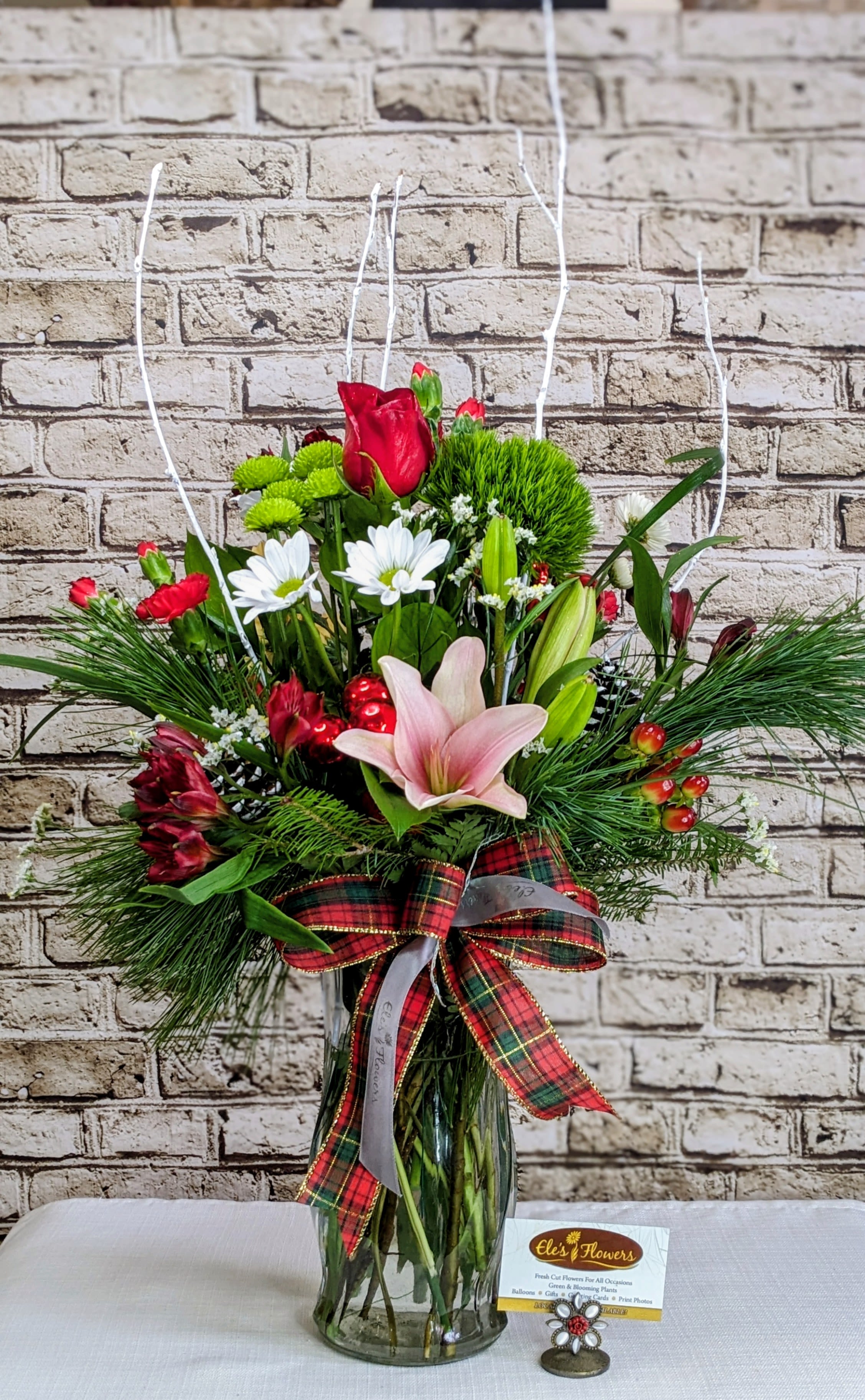 Holiday Elegance  - Bring classic holiday elegance into any space with this lush, winter-inspired floral arrangement designed to feel both festive and refined. Rising gracefully from a clear glass vase, this design features a striking mix of rich seasonal blooms, fresh evergreen accents, and whimsical curly willow branches that add height, movement, and drama.  Soft pink lilies and crisp white daisies provide a fresh, elegant contrast to deep red blooms and vibrant green textures, while glossy red accents and natural pine elements create warmth and seasonal charm. Finished with a timeless red-and-green plaid ribbon, this arrangement feels cozy, joyful, and unmistakably festive—perfect for celebrating the season in style.  Whether placed on a dining table, entryway, office reception, or gifted to someone special, this arrangement makes an unforgettable statement and fills the room with holiday spirit.  Ideal for holiday gifting, seasonal décor, or anyone who loves classic Christmas style with a fresh, modern twist.  Approximate Dimensions: 30"H x 20"W