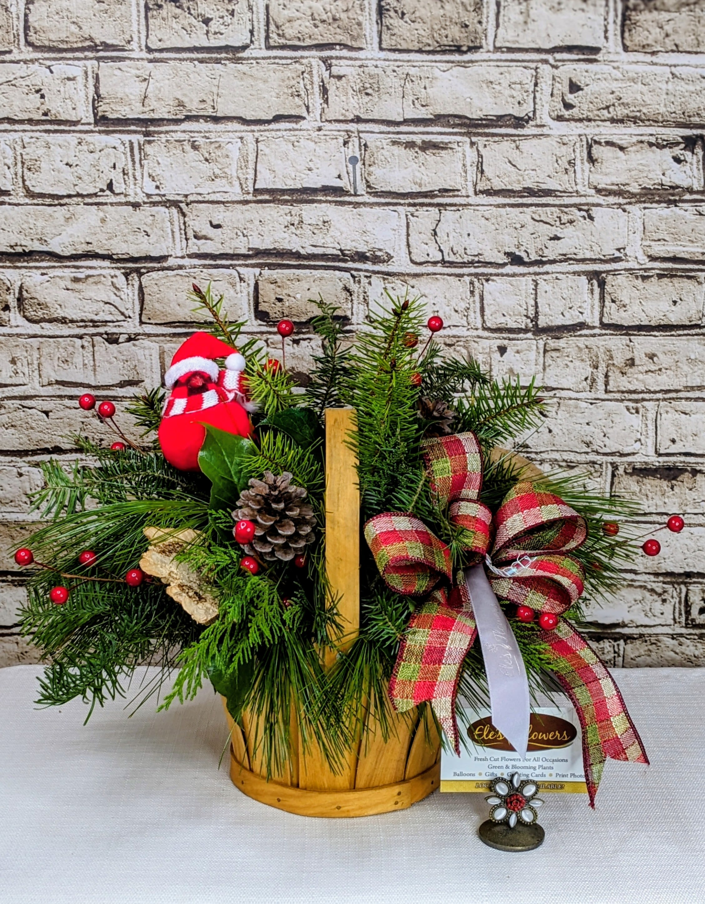 Winter's Charm - A charming holiday arrangement featuring a mix of lush, fragrant evergreens in a rustic wood chip basket. The design is enhanced with pops of vibrant red berries, scattered pinecones, and natural accents like mushrooms or birch for a festive touch. A delightful little cardinal perches gracefully within the greenery, adding a playful and cheerful note. This arrangement is crafted to last throughout the holiday season, bringing timeless elegance and a touch of the outdoors to any space.  The designs we create are custom and the blooms and containers will vary. We use the freshest products available to create the perfect floral combination for the recipient! If you have any special requests, please add those in the &quot;special instructions&quot; portion of your order to help us dial in your vision. Otherwise, our skilled designers, Brett &amp; Sara, will take it from here! Please note that the photos are examples only – floral selection, ribbon and embellishments will vary.  Approximate Dimensions: 14H x 14&quot;W