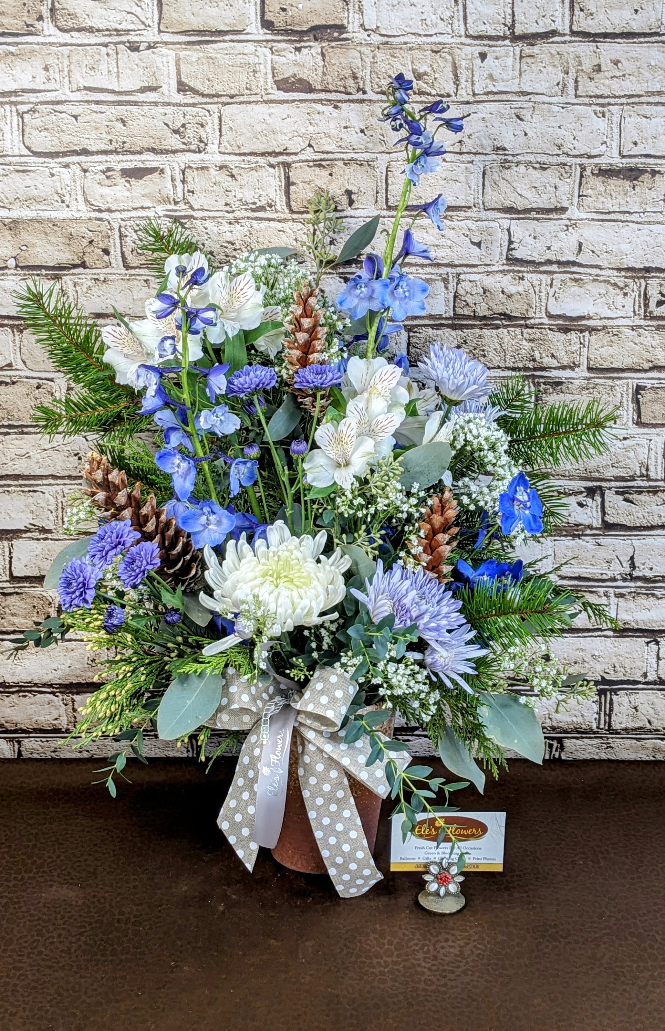 Winter Wonderland - A serene holiday arrangement featuring a harmonious blend of soft blues and crisp whites, evoking a winter wonderland. Fragrant evergreens provide a lush and aromatic base, while rustic pinecones add natural texture and warmth. This elegant arrangement brings a touch of frosty beauty and tranquility to any holiday décor.  Blooms and container may vary from photo.  Approximate Dimensions: 25"H x 19"W