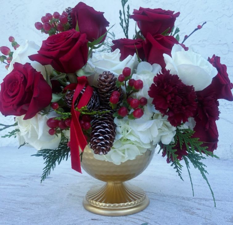 Crimson Noël - Rich crimson roses rise above soft clouds of white hydrangea, accented with deep burgundy carnations, winter berries, and natural pinecones for a timeless holiday statement. Designed in an antique-inspired gold pedestal vessel, this arrangement blends traditional Christmas warmth with refined elegance.  Lush evergreens and subtle festive details create depth and texture, making this piece perfect for holiday gifting, formal gatherings, or a classic seasonal centerpiece. Sophisticated yet welcoming, Crimson Noël brings the beauty of winter indoors with understated luxury.  Available for a limited time during the holiday season.