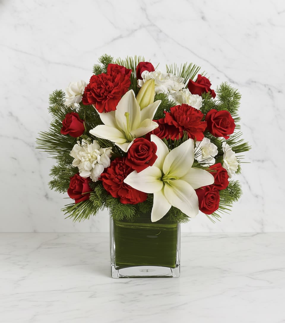 Christmas Lily Delight - Celebrate the season with this elegant holiday arrangement featuring radiant white lilies, classic red roses, cheerful red and white carnations, and lush winter greenery. Designed in a modern glass cube wrapped with a fresh green leaf lining, this compact yet striking bouquet brings a perfect balance of festive color and contemporary style. Ideal for holiday gifting, office décor, or brightening your home with a touch of winter charm, this arrangement adds warmth and beauty to any space.