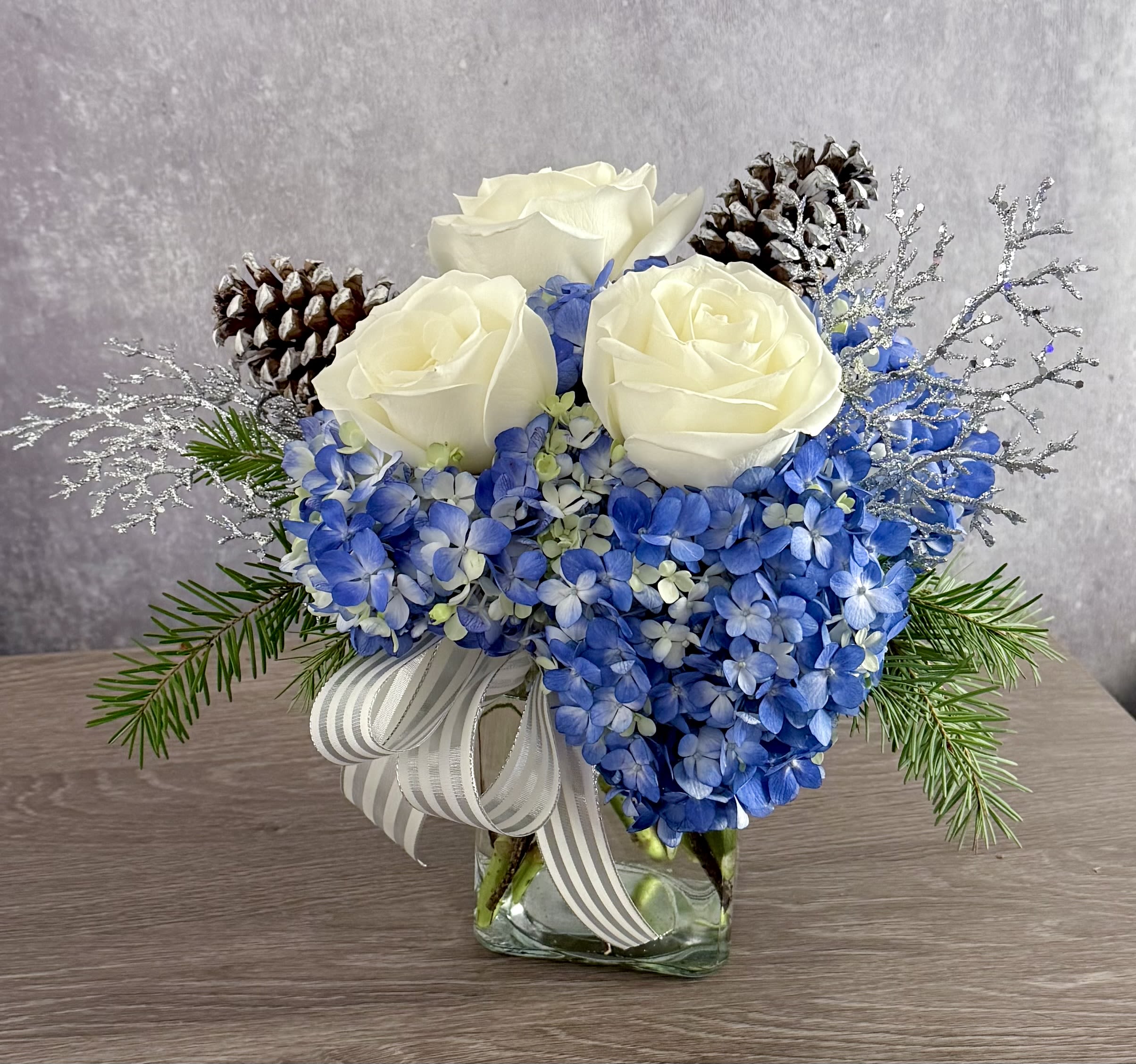 Winter Splendor  - A clear glass vase with a cluster of blue hydrangeas, 3 white roses, pine cones and silver sparkle picks. Soft and pretty for winter! 