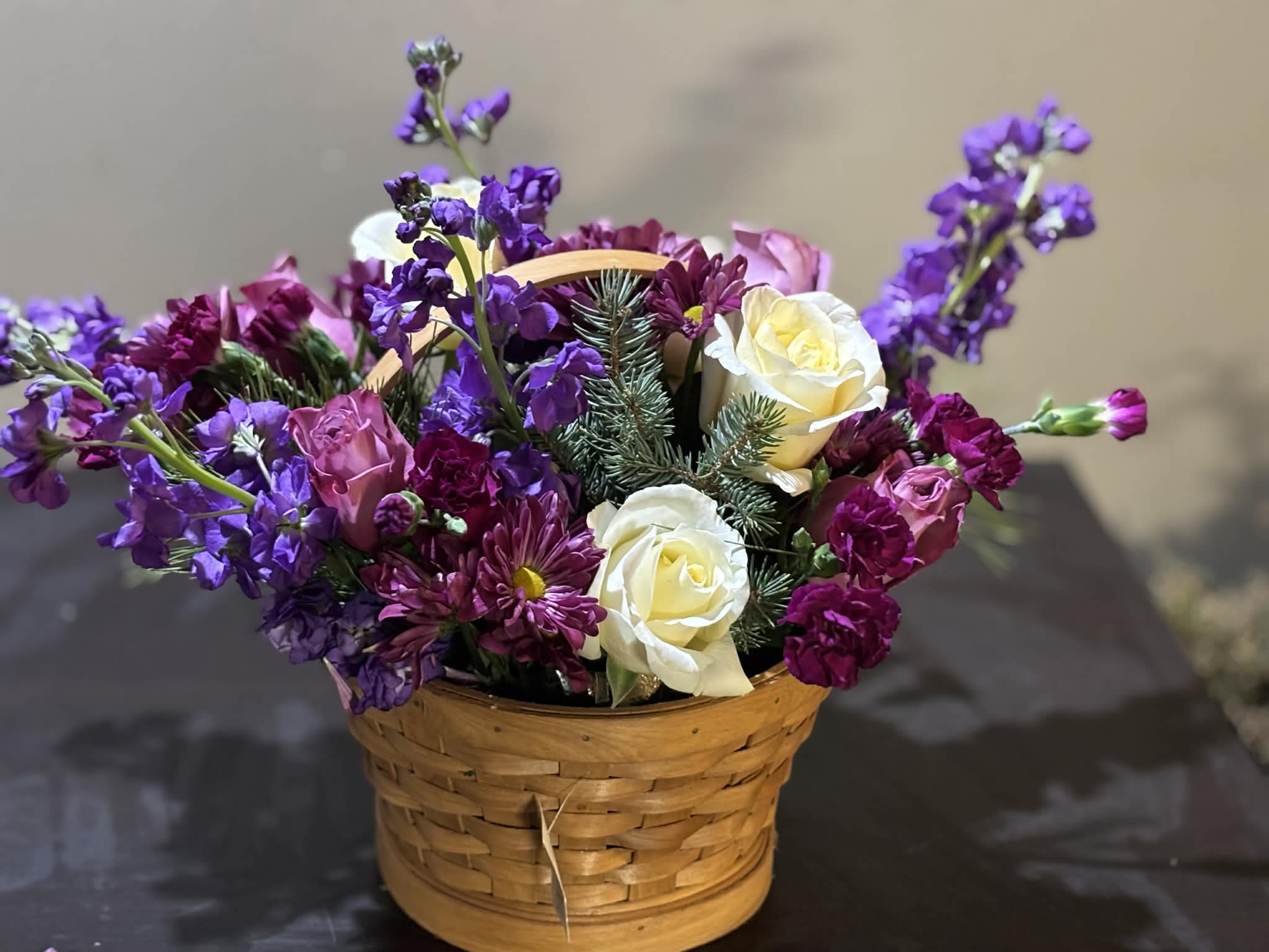 Plum &amp; Ivory Garden Basket - A charming woven basket overflowing with rich plum, violet, and ivory blooms. Creamy white roses are nestled among deep purple chrysanthemums and delicate lavender stock, accented with fresh evergreen sprigs. This arrangement feels warm and elegant, blending classic florals with a cozy, handcrafted touch—perfect for winter gifting or a welcoming centerpiece.