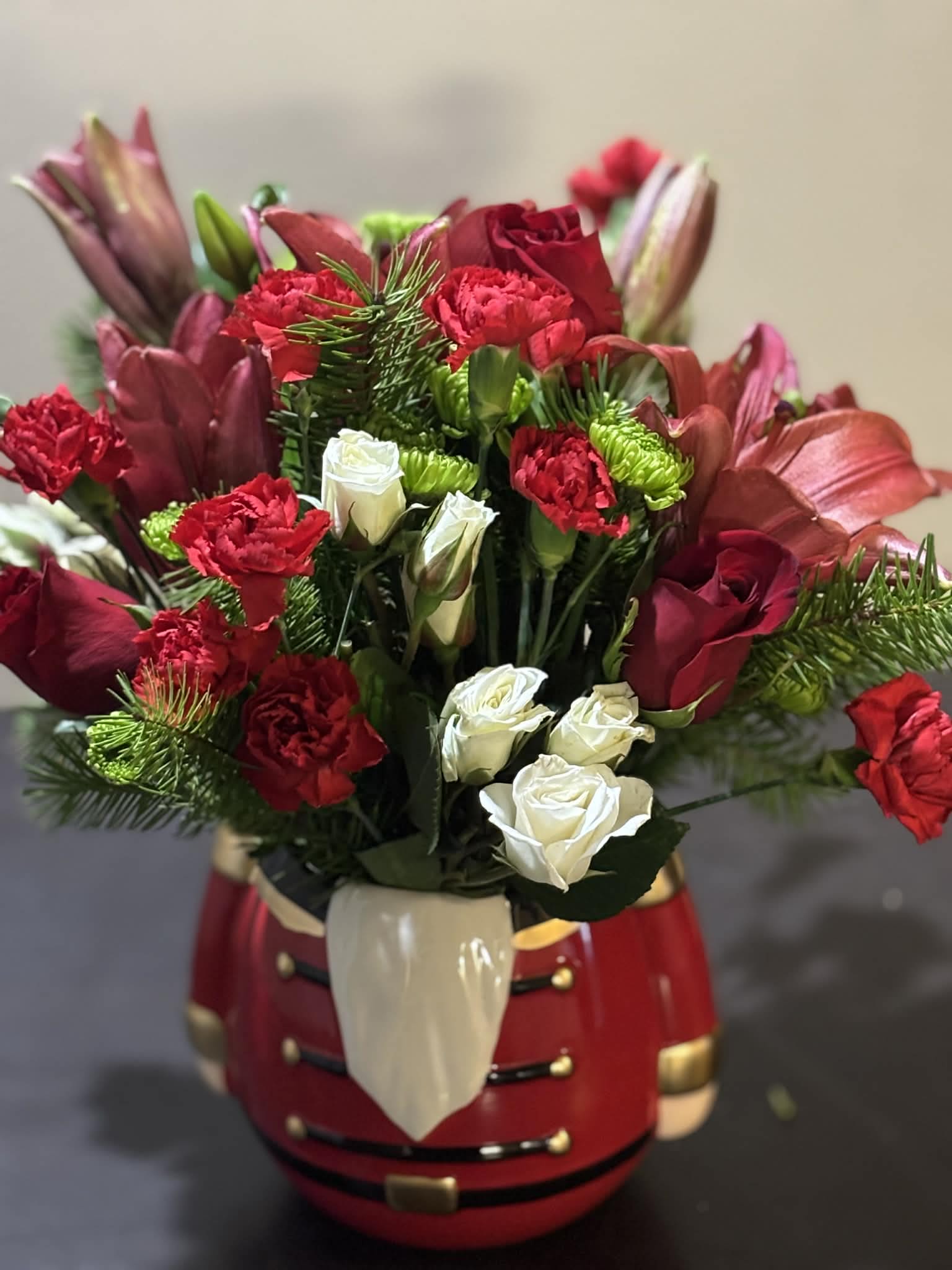 Festive Holiday Harmony - This lush holiday arrangement combines deep red roses, white spray roses, red carnations, and vibrant greenery, all beautifully arranged in a festive nutcracker container. Full of texture and seasonal spirit, it’s ideal for Christmas celebrations, gifts, or adding a bold holiday statement to any space.