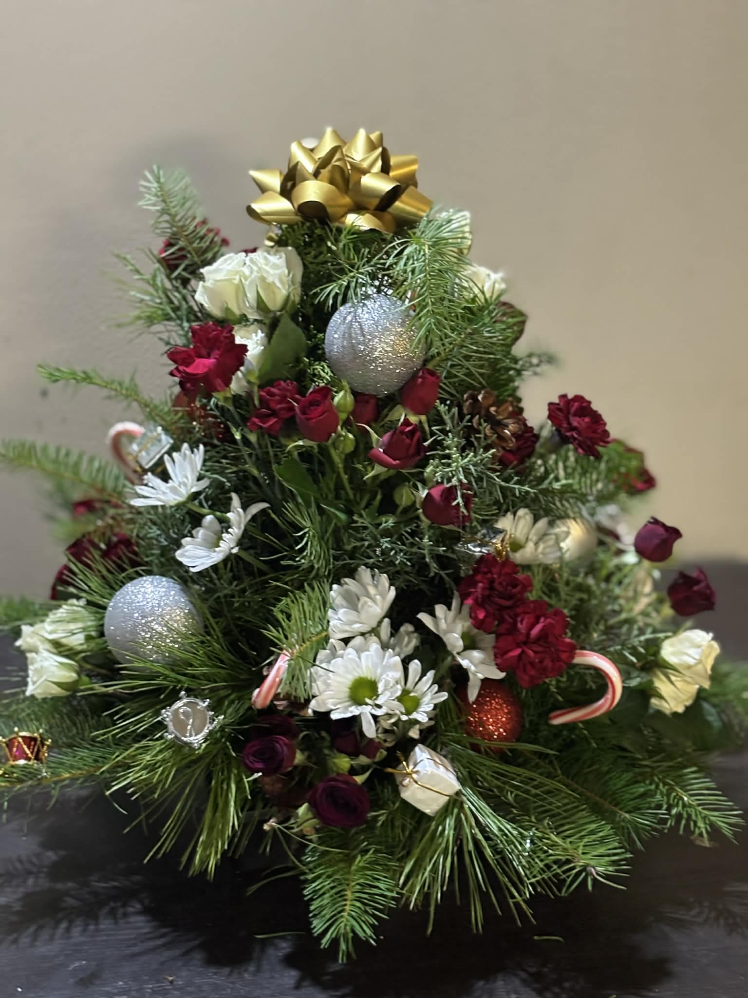 Golden Christmas Tree Delight - A charming Christmas tree–shaped arrangement filled with fresh evergreen branches, rich red spray roses, white daisies, and elegant white roses. Accented with festive ornaments, candy canes, pinecones, and topped with a beautiful gold bow, this design captures the magic and warmth of the holiday season. Perfect as a centerpiece, gift, or festive home décor piece.