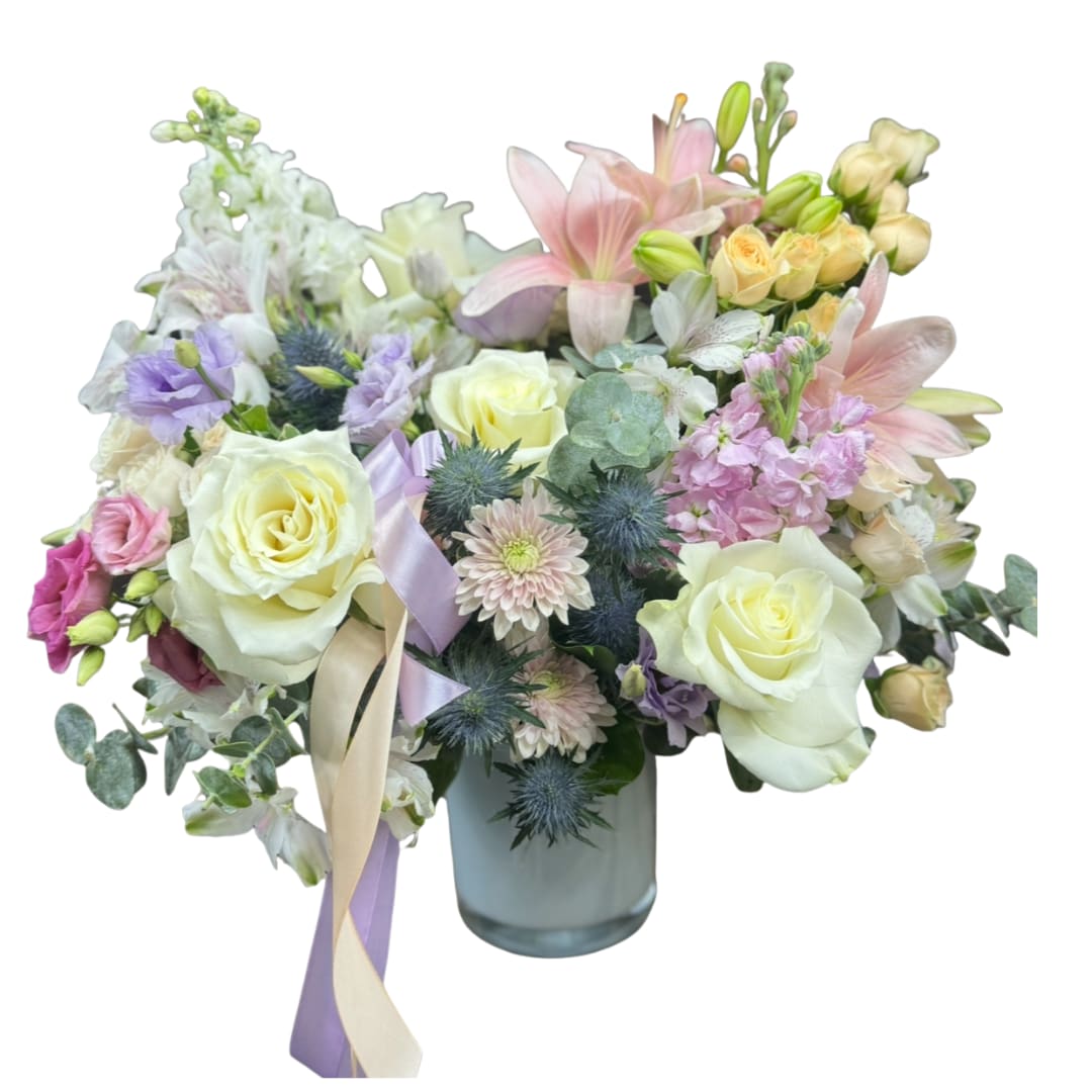 Daydream - A dreamy pastel mix of ivory roses, blush lilies, buttery spray roses, and lavender blooms, finished with blue thistle + eucalyptus for texture. Designed in a clean glass vase with soft ribbon  sweet, airy, and super giftable.