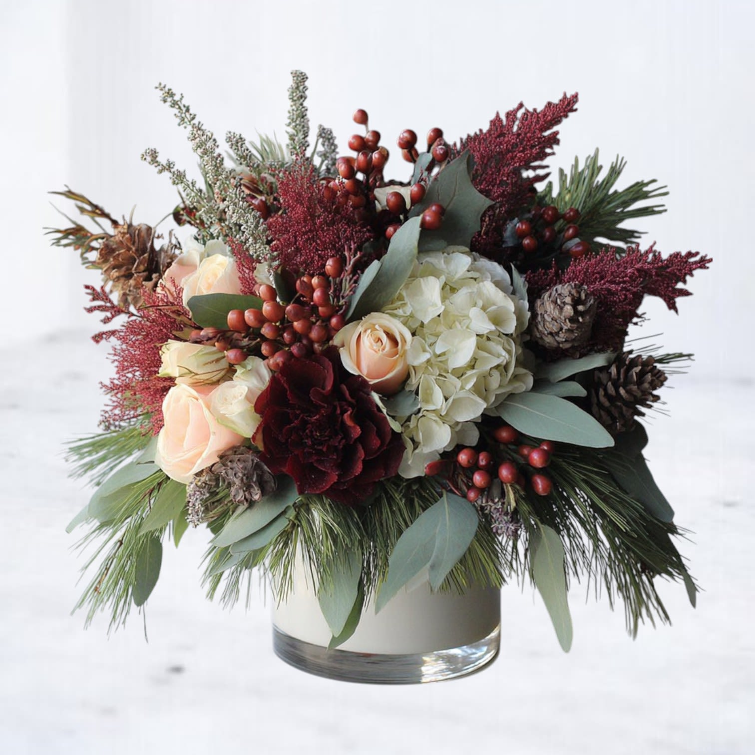 Hearthwood Noel - Warm, textural, and richly seasonal, Hearthwood Noel captures the feeling of a winter gathering by the fire. This garden-style holiday arrangement blends creamy roses, soft ivory hydrangea, and deep wine-toned blooms with layers of fresh evergreen, eucalyptus, pinecones, and clusters of winter berries.  Designed with natural movement and a full, lush silhouette, this arrangement balances rustic charm with refined elegance. Burgundy accents and woodsy textures create depth and contrast, while neutral florals soften the palette for a timeless holiday look. Styled in a classic glass vessel, Hearthwood Noel is perfect for winter entertaining, seasonal gifting, or adding warmth to holiday décor.