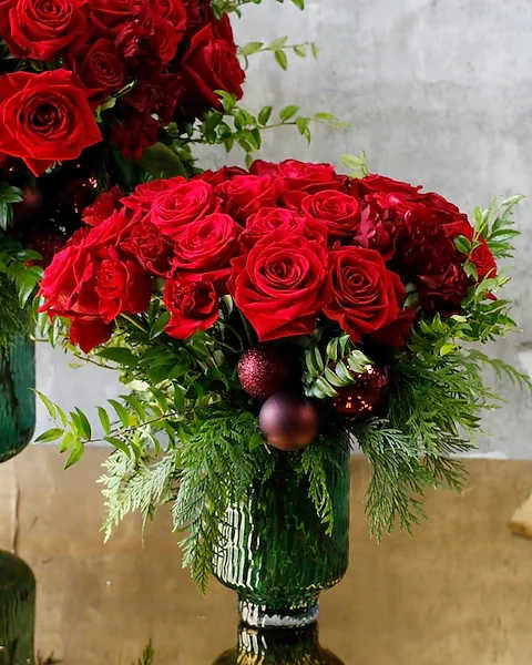 Christmas Couture - Vibrant Red roses and Shooting Star spray roses intertwine with lush huck and fragrant incense cedar, creating a bold yet elegant composition. Set against a textured emerald vase, this arrangement radiates a rich, festive allure, accentuated by clusters of wine ornaments that evoke the warmth of holiday gatherings. The Christmas Roses embodies couture sophistication, transforming any space into a luxurious winter wonderland that captures the enchanting spirit of the season. Ideal size for making a statement in the entry-way or on a buffet table.