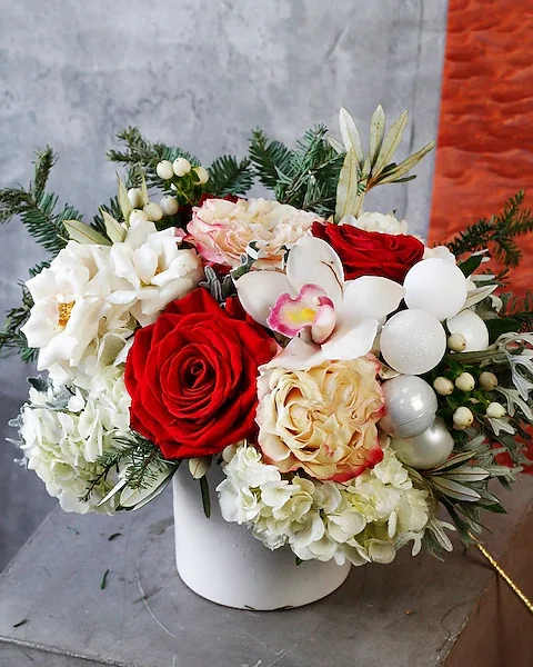 Candy Cane Christmas - A whimsical blend of textures and colors defines Candy Cane Christmas, where the soft lace leaf dusty miller and white hydrangea create a luxurious backdrop for striking white cymbidium blooms and rosy accents. Vibrant Red roses, Candy roses, and Sedna spray roses bring a festive flair, while frosted pinecones and sprigs of noble fir add a touch of winter’s charm. Nestled within a white ceramic cylinder vase, this arrangement captures the enchanting spirit of the season, evoking the joy and wonder of a magical holiday carousel. Ideal size for a kitchen table or island, coffee table, entry-way or large desktop.