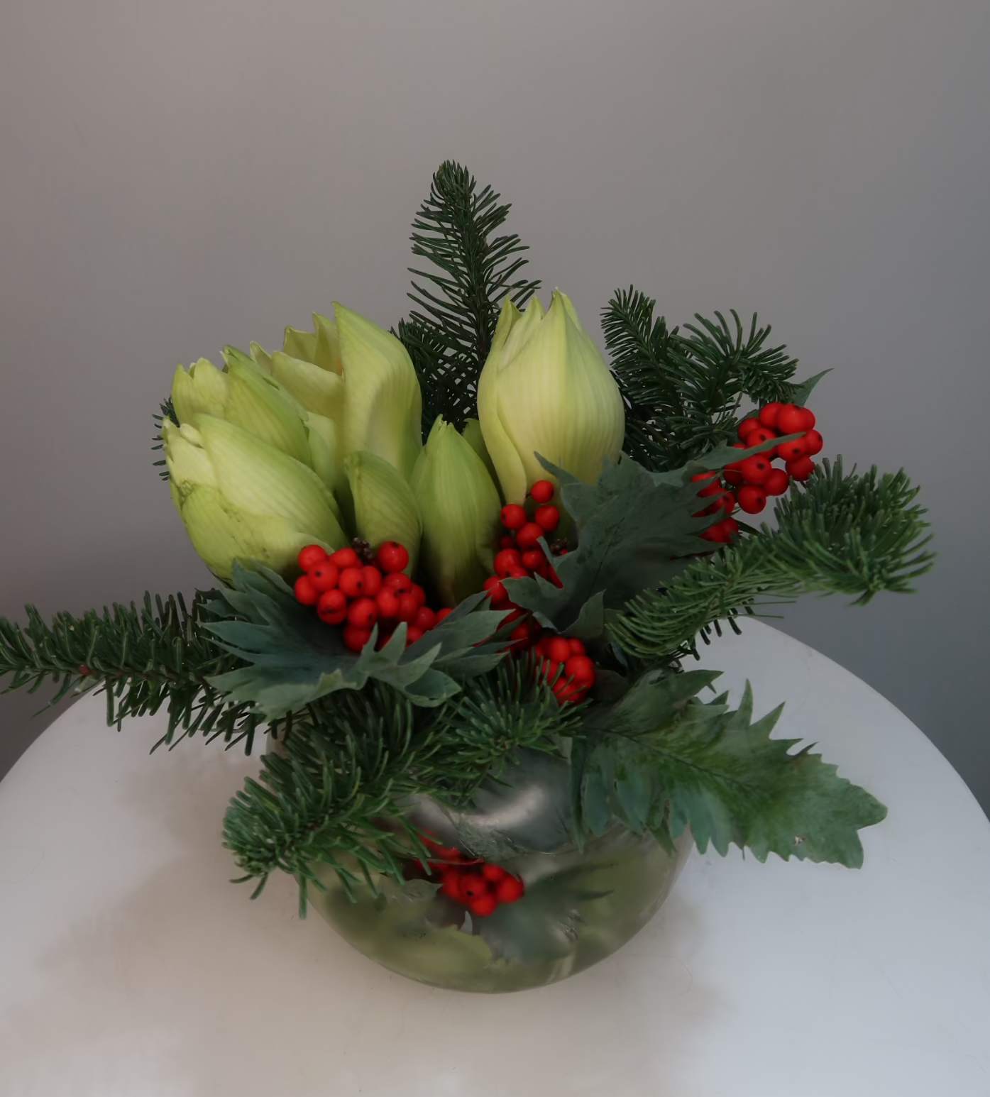 Winter Amaryllis Berry Garden - A festive winter arrangement featuring fresh green amaryllis buds, fragrant evergreen branches, and vibrant red berries arranged in a modern glass vase. This seasonal design brings natural elegance and holiday warmth to any space, making it perfect for Christmas decor, winter centerpieces, or thoughtful seasonal gifting. Designed to bloom beautifully over time, this arrangement adds a fresh and timeless holiday touch to homes and offices.