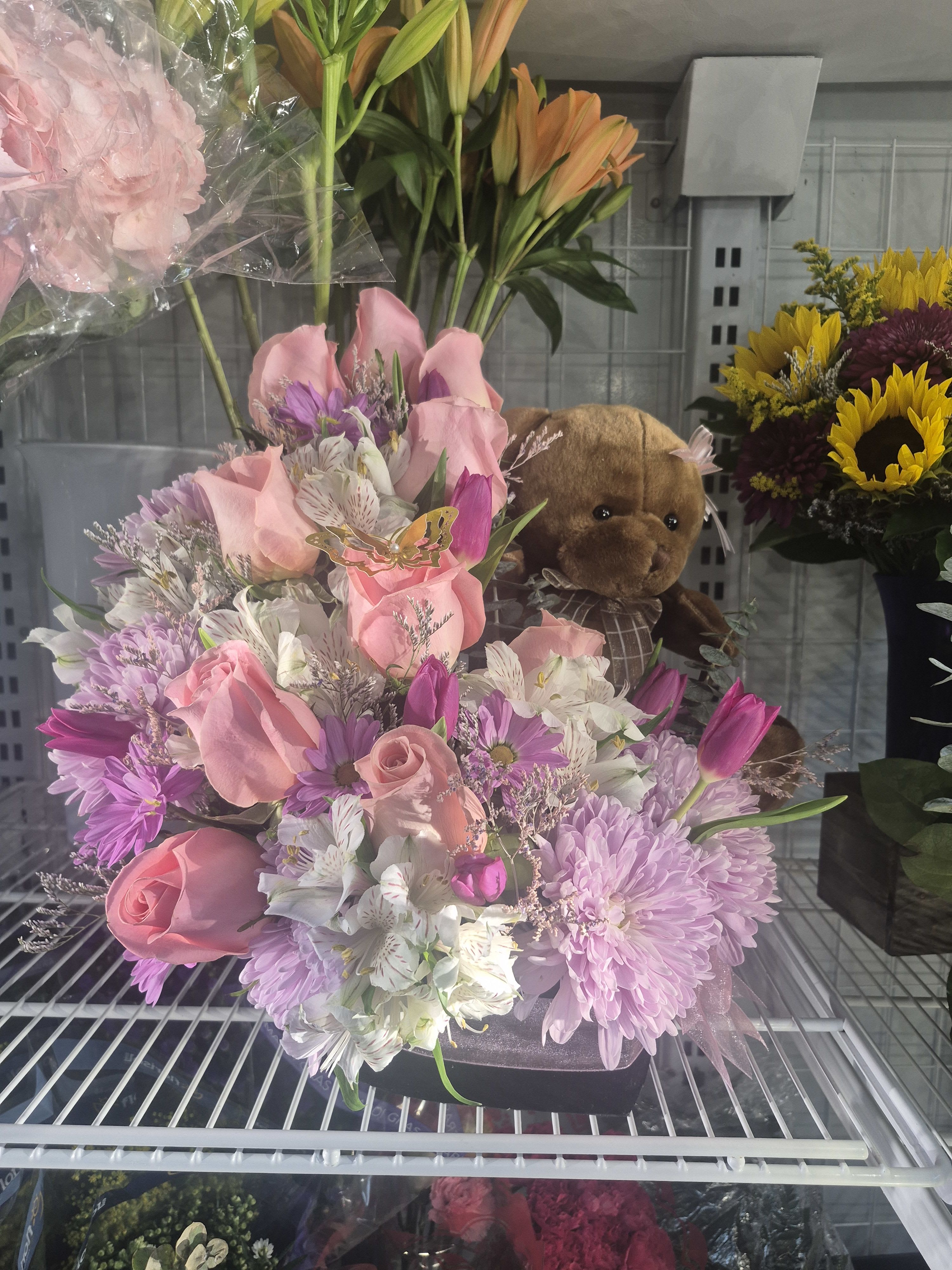Teddy Bear - This is a beautiful and very sweet floral arrangement. It features a mix of soft pink and purple flowers, including roses and tulips ,arranged to look full and lush. The best part is the cute teddy bear nestled right in the middle, giving the whole bouquet a warm and cozy feeling. It’s the perfect way to send a hugs and flowers message.