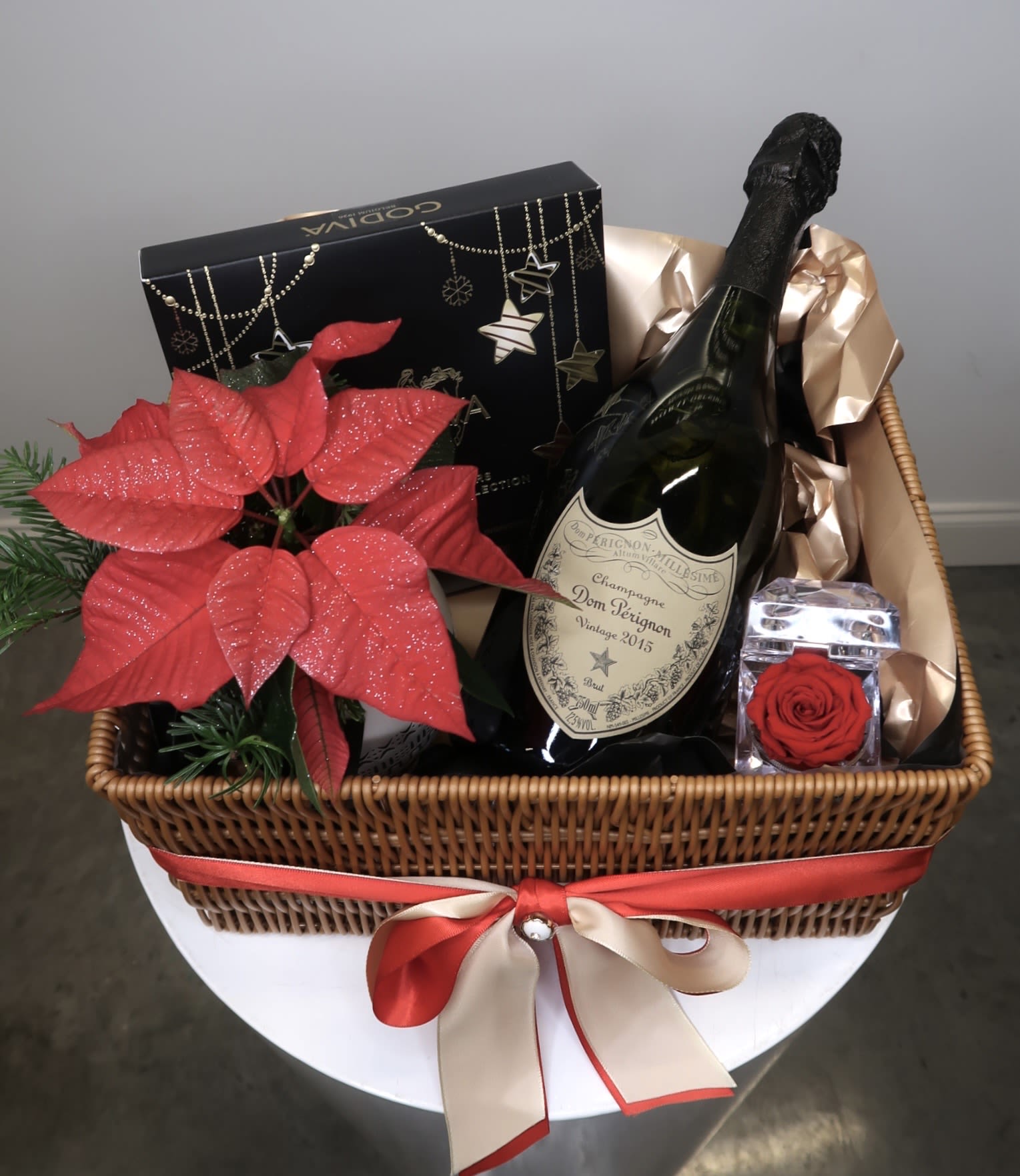 Holiday Dom Pérignon Celebration Basket - An elegant holiday gift basket featuring a premium bottle of Dom Pérignon champagne, a festive red poinsettia plant, gourmet chocolates, and a preserved red rose accent, beautifully arranged in a woven keepsake basket with luxury ribbon detailing. This sophisticated holiday gift is perfect for Christmas celebrations, corporate gifting, anniversaries, and special occasions. Designed to impress, this curated set blends floral elegance with luxury indulgence for a truly unforgettable presentation.