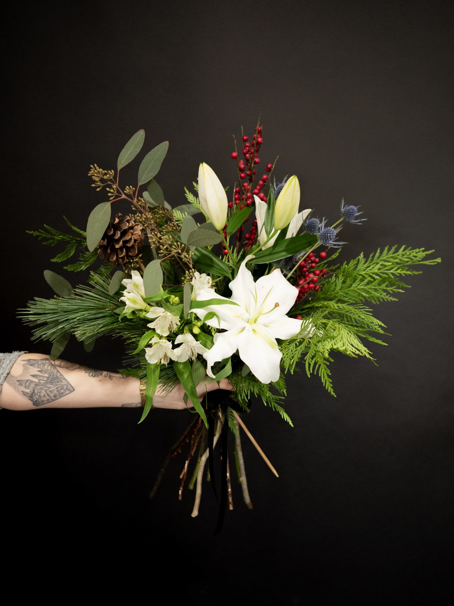 Designer's  Choice, Winter Elegance - Hand tied Bouquet - Celebrate the magic of the season with our Winter Elegance Bouquet. This hand-tied arrangement features pure white lilies and delicate alstroemeria, framed by fresh evergreens and eucalyptus for a lush, festive feel. Accents of natural pinecones, red ilex, and blue eryngium add vibrant holiday touches, making it perfect for any winter occasion.  Whether it's for a gift or to adorn your home, this bouquet radiates seasonal charm. We offer customization options to personalize your arrangement with extra blooms or unique accents tailored to your preferences.
