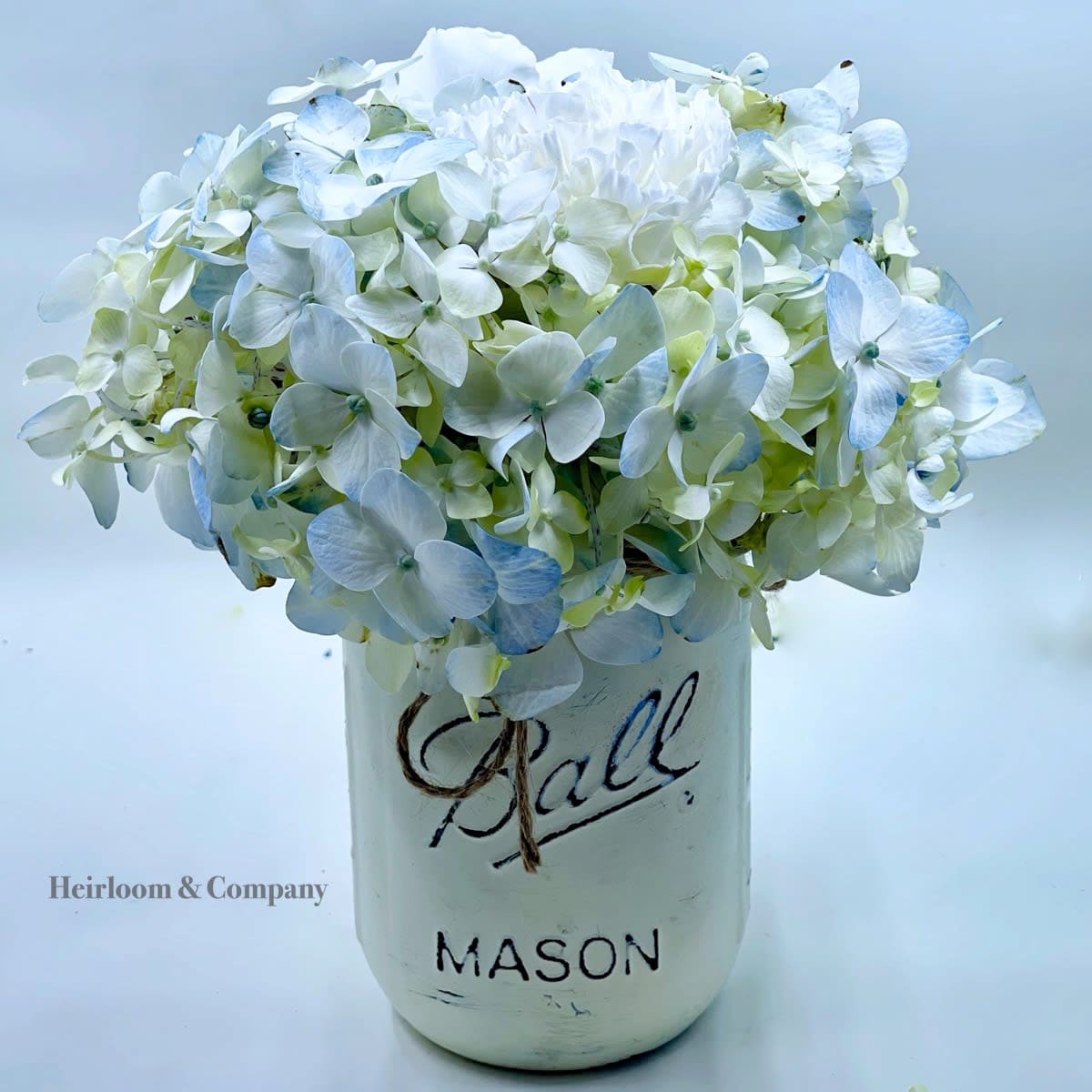 Blue Haze - The freshest and loveliest sky blue hydrangea sprinkled with white caranations and mini ivory spray roses, all presented to you in a hand painted ivory mason jar with a twine bow!