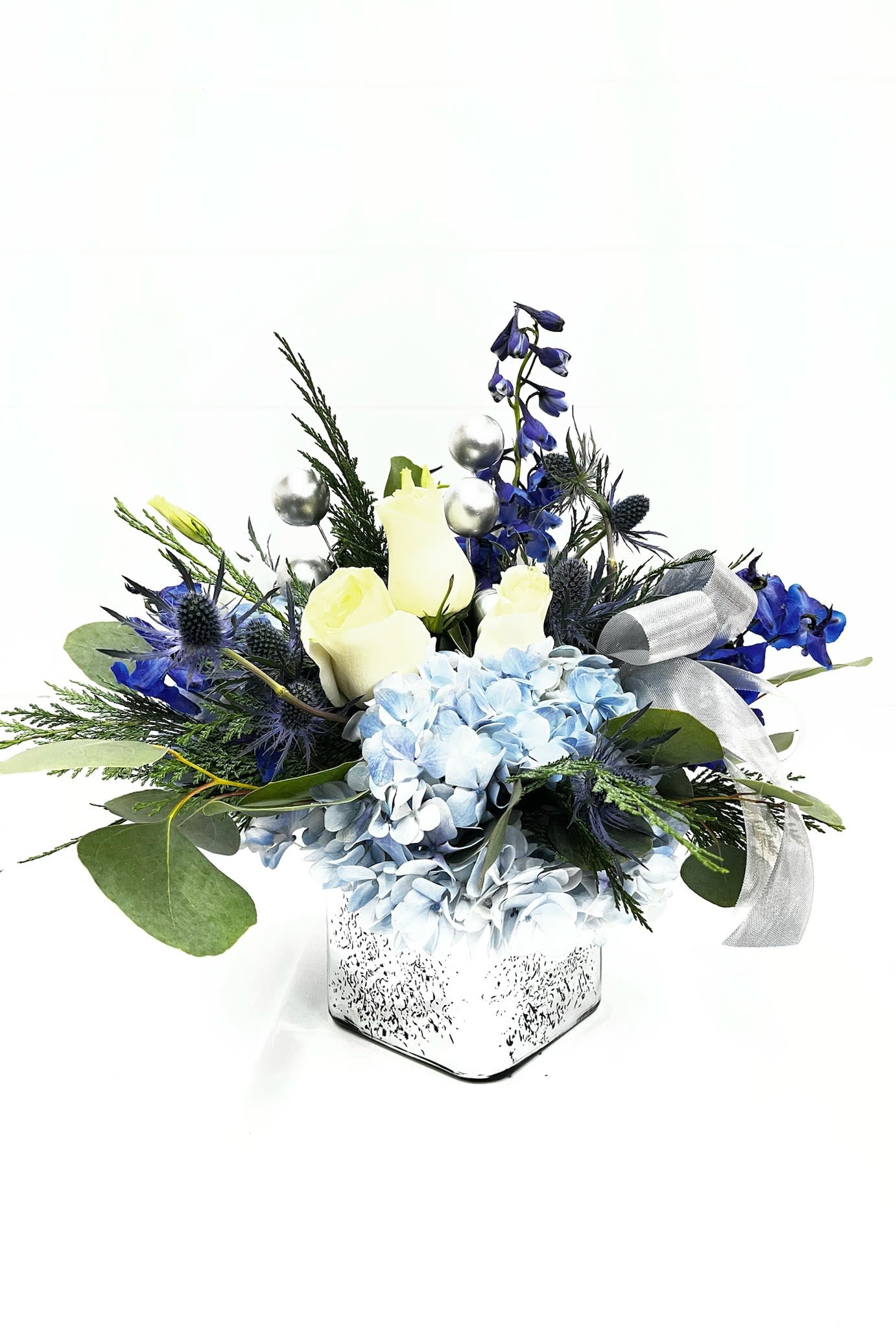 Winter's Calm - Winter's Calm Bouquet  A cool winter arrangement featuring blue hydrangea and delphinium, creamy white roses, festive Christmas greens, and silver accents, designed in a reflective silver cube with a silver bow. Perfect for both Hanukkah and Christmas, this elegant piece works beautifully as a holiday centerpiece, entryway accent, or office display, adding a refined seasonal touch to any space.