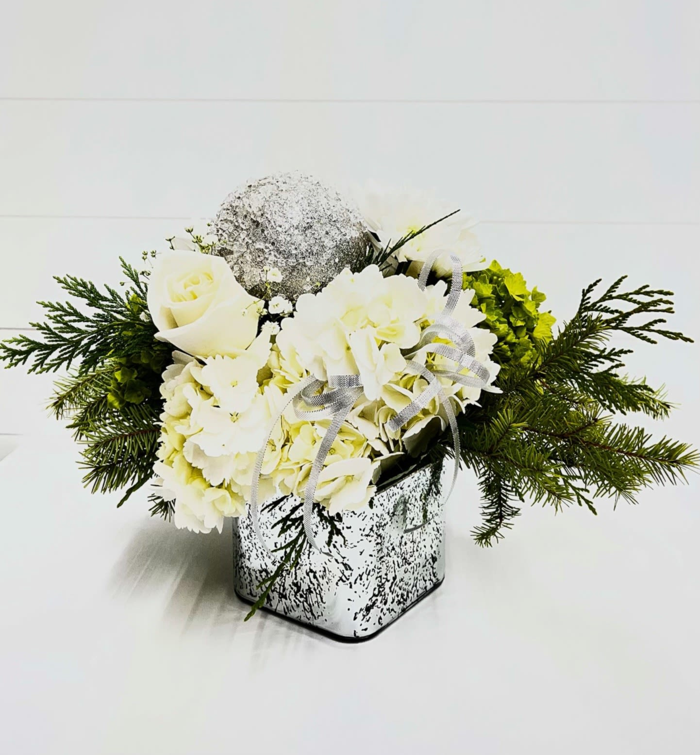 Silver Tidings - Fresh white hydrangeas and roses paired with winter evergreens, finished with a silver ribbon and shimmering silver ornament. Arranged in a reflective glass cube, this piece offers a clean, elegant look perfect for holiday décor, winter centerpieces, or seasonal gifting.