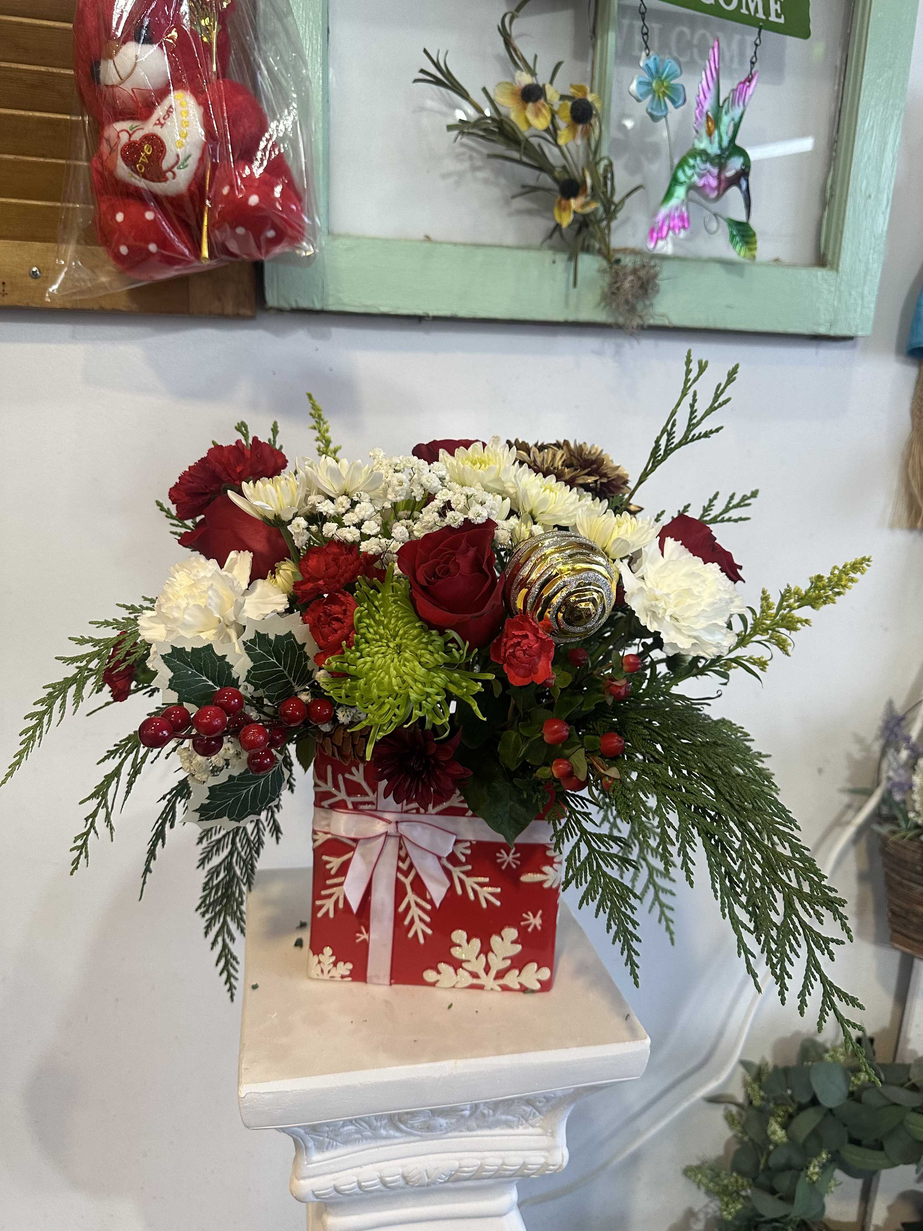 Holiday Suprise Box  - This floral arrangement is atop a decorative pedestal. The bouquet is set in a square container designed like a wrapped gift box, adorned with white snowflakes and a ribbon against a red background, enhancing the holiday theme. It includes red and white flowers like roses and possibly carnations, with lush green elements like ferns and potentially chrysanthemums. Bright red berries and a gold-striped ornament nestled among the flowers add a touch of elegance.