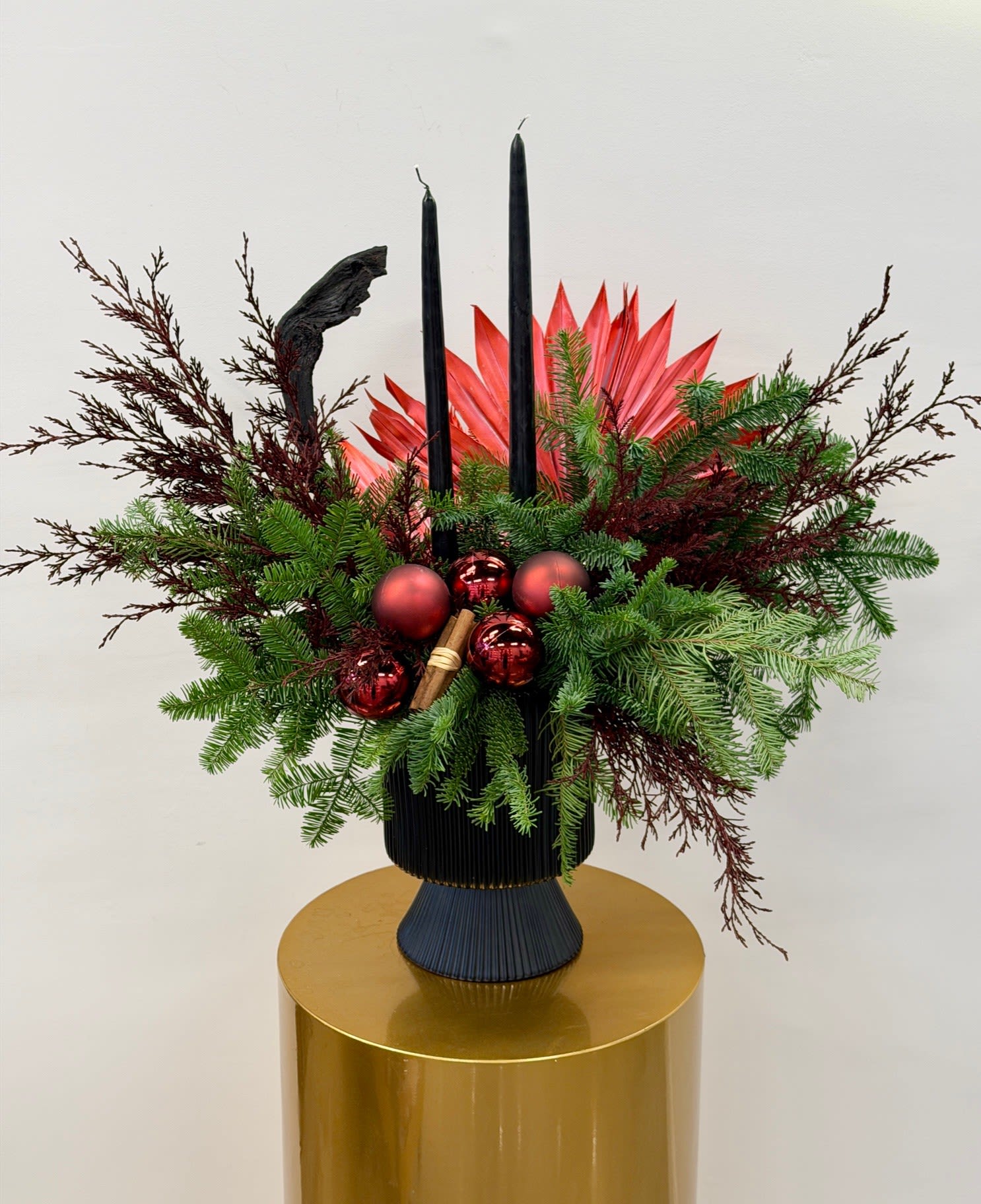 Midnight Crimson™ - A striking holiday centerpiece that blends drama and elegance, Midnight Crimson features lush seasonal evergreens layered with deep red accents, sculptural foliage, and sleek black taper candles. Finished with rich ornaments and textural details, this arrangement evokes a modern winter soirée with a hint of moody glamour.  Designed to make a statement, it’s perfect for holiday tables, entryways, or elevated celebrations where bold design meets festive sophistication.