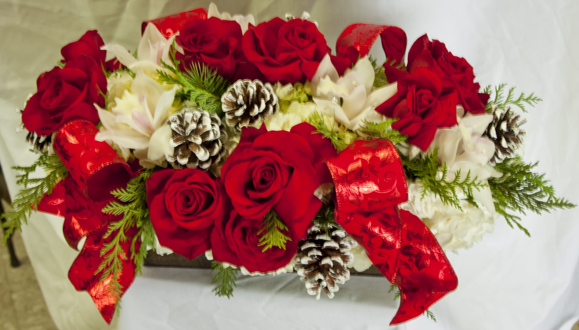 HaleyCenterpiece - Long and low centerpiece with assortment of red roses, white hydrangeas, white cymbidium orchids, pine cones and beautiful Christmas ribbon.