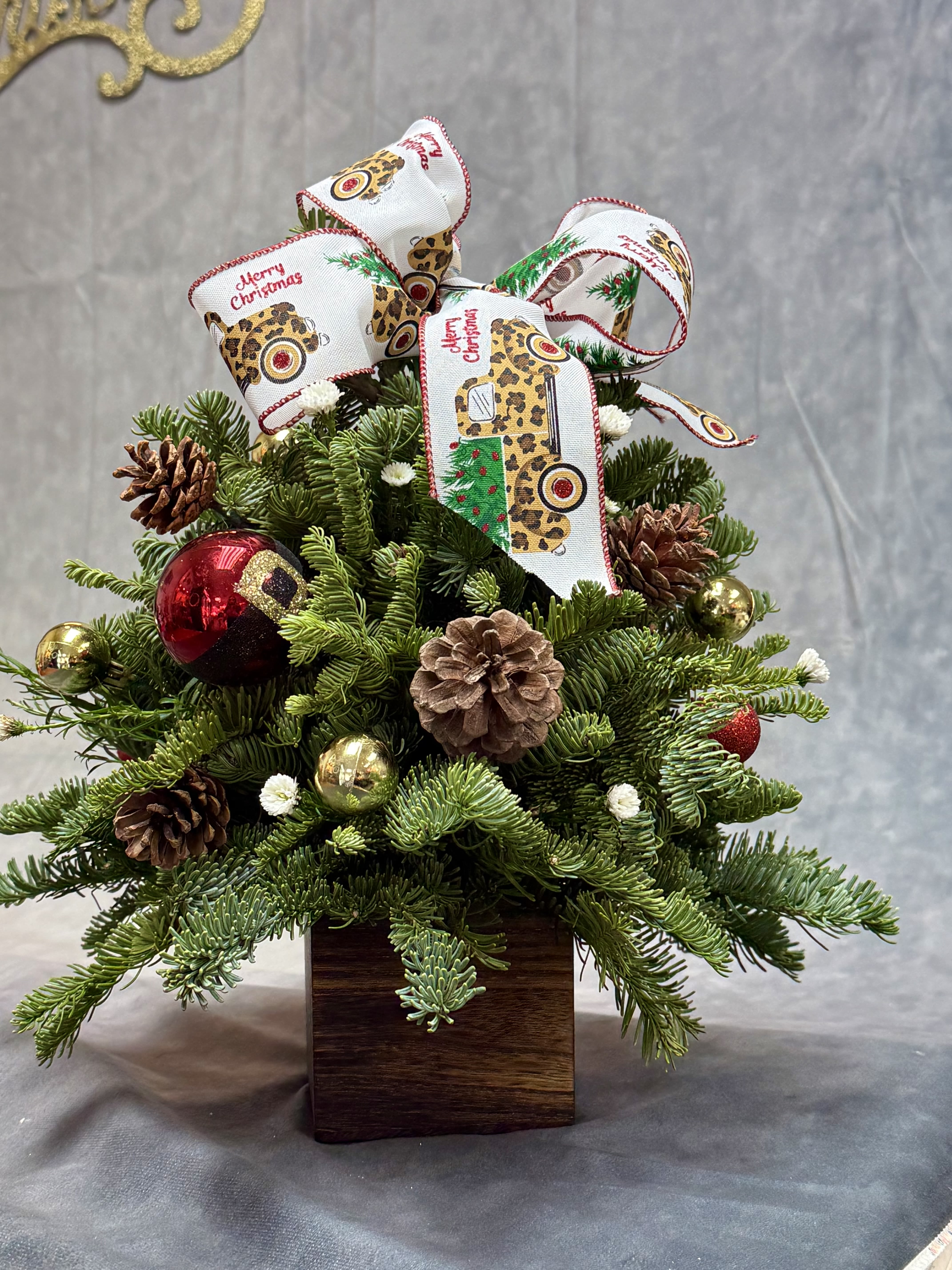 Christmas Tree Box - A charming Christmas tree box designed with fresh evergreen greenery, pinecones, festive ornaments, and a joyful holiday bow—bringing the warmth and magic of the season into any space.it’s 15”H x 10” W 