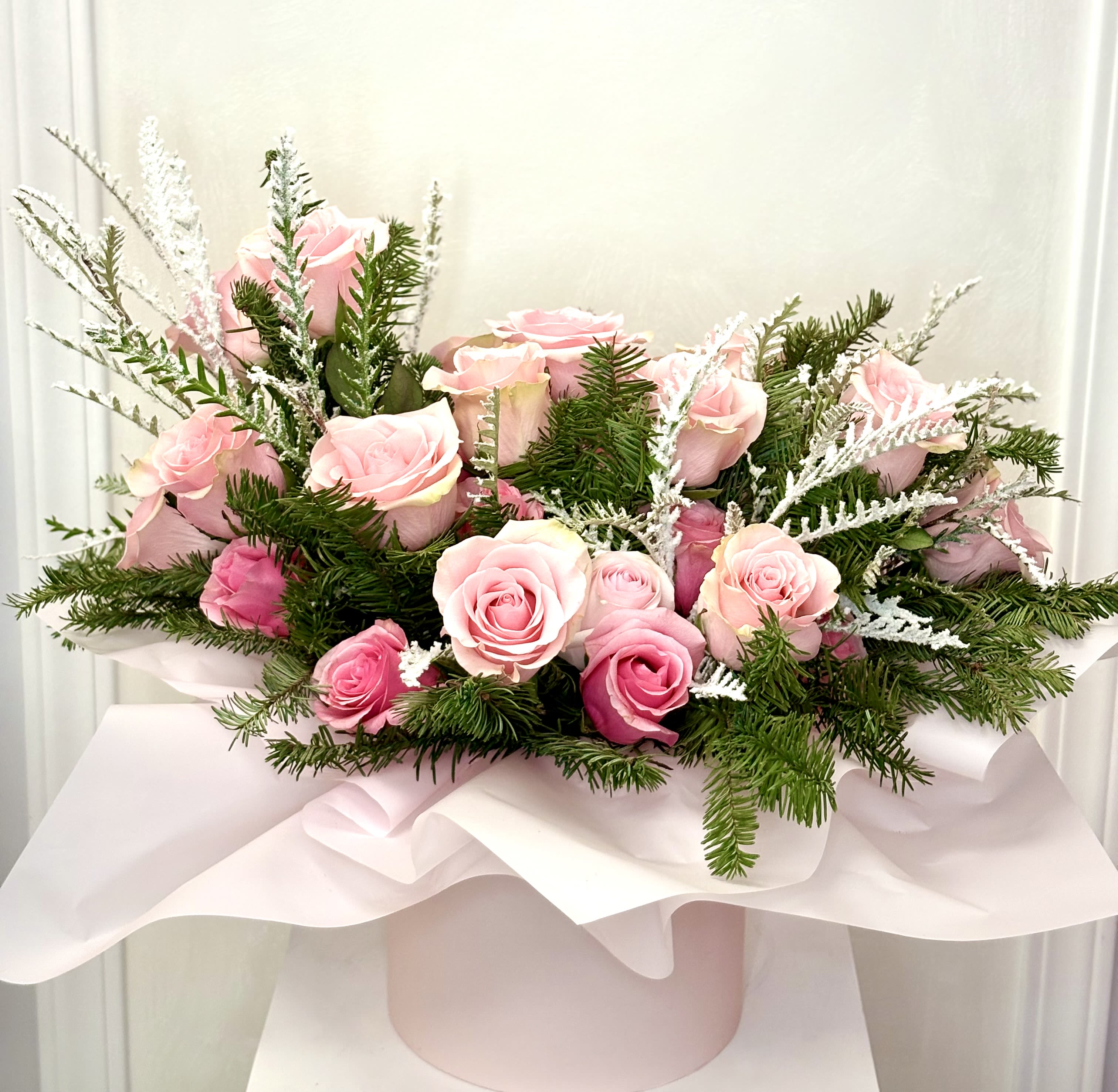 Blushing Winter Grace By VLM Flowers - A soft and elegant arrangement of blush and pink roses nestled among fresh winter evergreens and delicate frosted accents. Designed in a gentle, low profile with flowing white wrap and a pastel pink base, this piece captures a romantic winter feel—perfect for heartfelt gifts, seasonal décor, or celebrating tender moments with timeless beauty.
