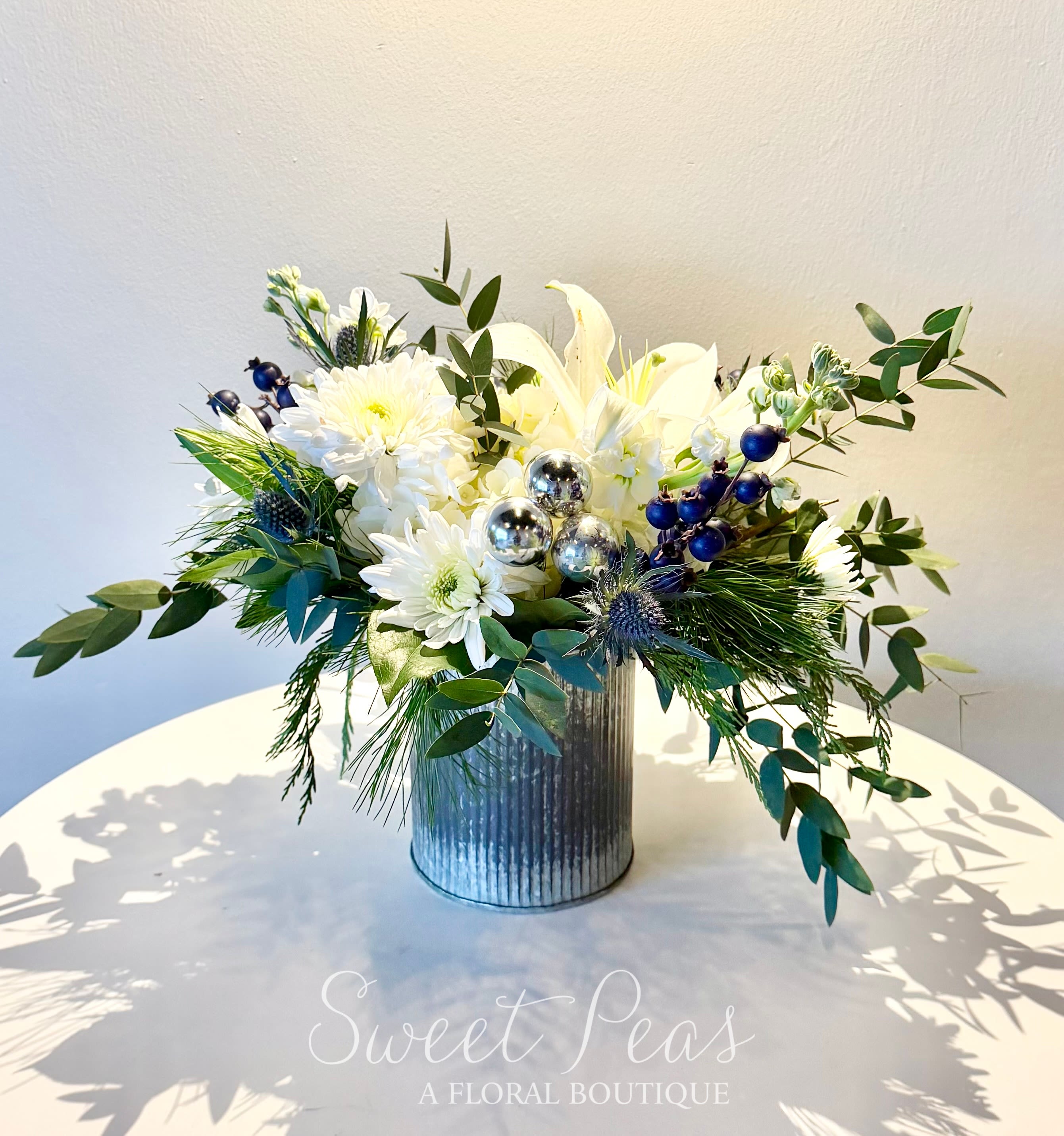 Silver Solstice - Elegant and luminous, Silver Solstice captures the quiet sparkle of the winter season. Featuring pristine white blooms layered with soft winter greenery, textural accents, and shimmering silver ornaments, this arrangement offers a fresh, modern take on holiday florals. Cool blue berries and sculptural elements add depth and contrast, all thoughtfully designed in a contemporary ribbed container for a polished finish.  Perfect for winter celebrations, holiday décor, sympathy, or an elevated seasonal gift, Silver Solstice brings a sense of calm beauty and refined sparkle to any space.  Details:  Color palette: White, silver, soft blue, and fresh green  Features seasonal white blooms, berries, and evergreens  Designed in a modern decorative container  Handcrafted; each arrangement is one of a kind