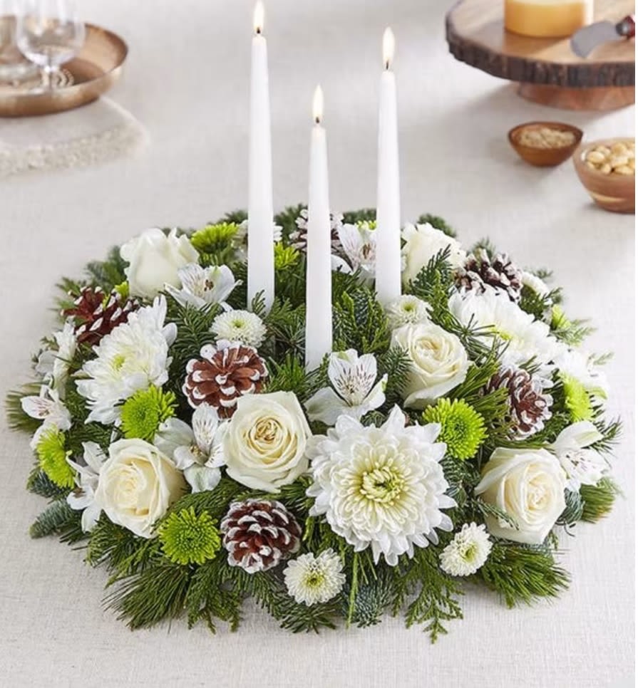 Winter's Charm™ Centerpiece -  Winter has a charm all its own, which we’ve brought to light in our brand-new seasonal centerpiece. Delightfully designed with a gathering of white blooms, snowy-tipped pinecones and fragrant greenery, this rustic, round-shaped arrangement is finished off with white taper candles, bringing a warmth and glow to their home.