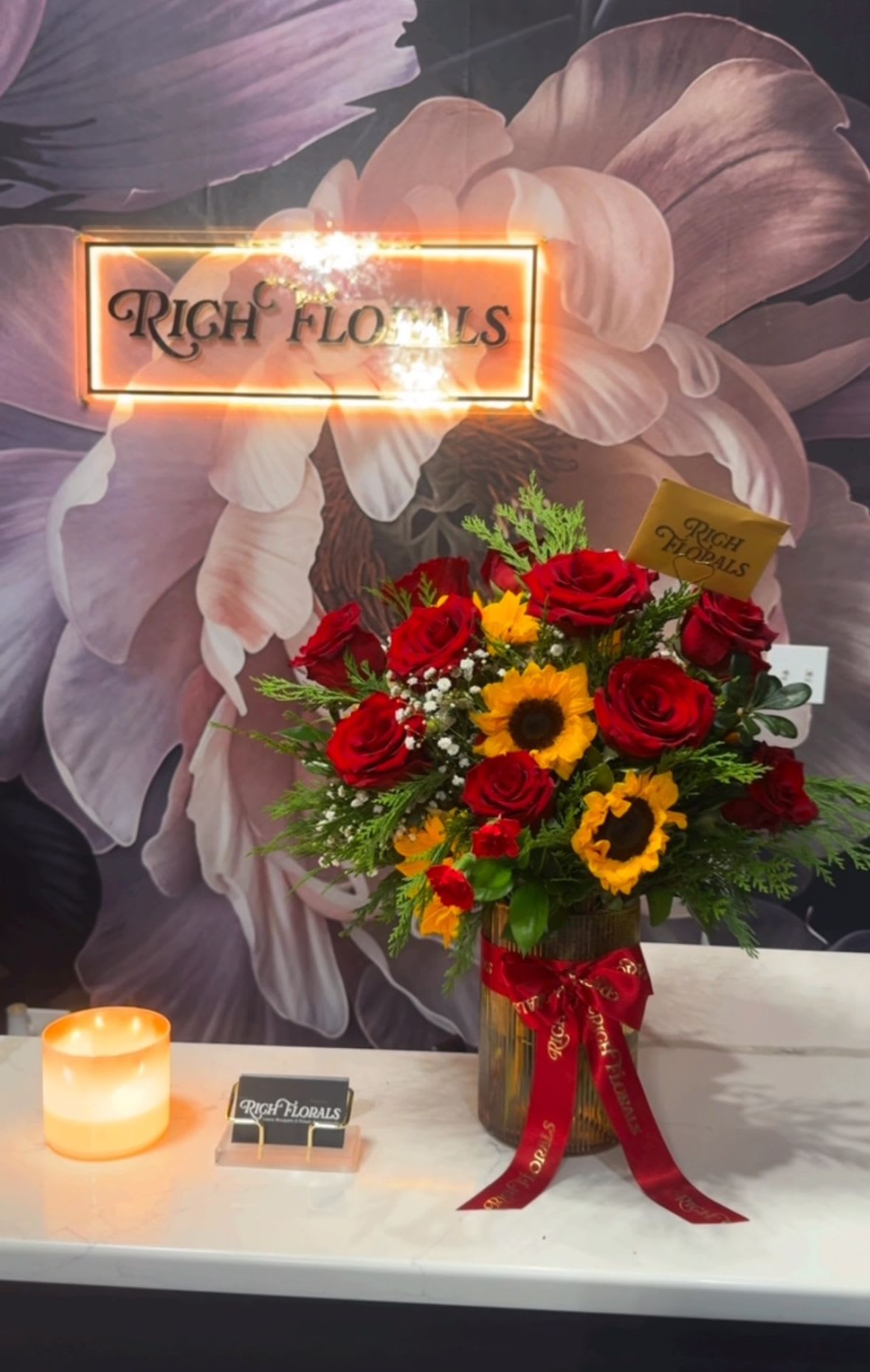 Crimson Son Vase Arrangement - Fifteen red roses and half a dozen sunflowers. The mix is classic, the execution is modern. Deep reds against bright golds with greenery that actually complements instead of just filling space. Comes in a glass vase with our signature ribbon.