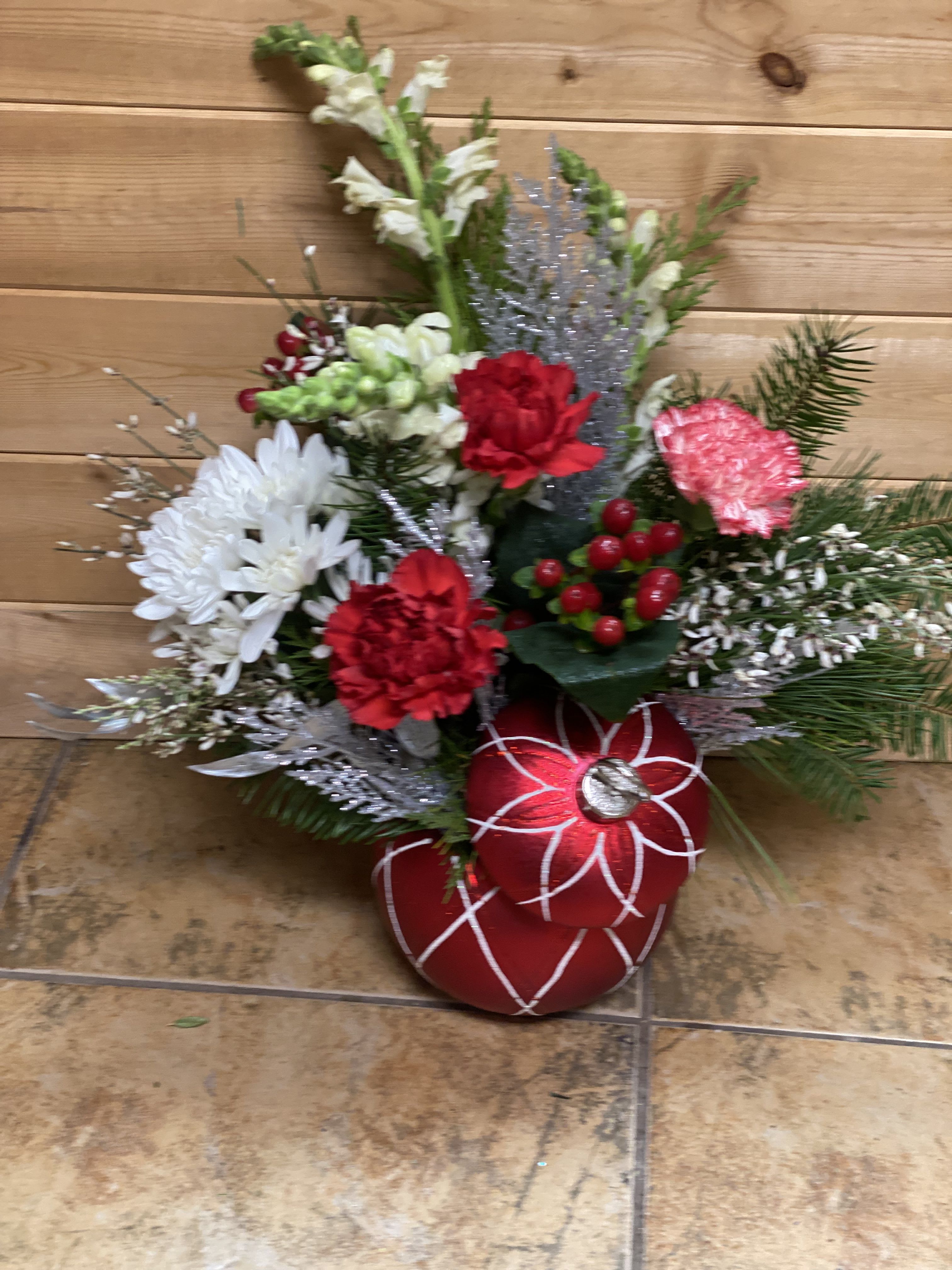 Crimson Cheer Evergreen Arrangement - Bring festive warmth to any space with this charming holiday arrangement. Crimson Cheer features rich red carnations, crisp white mums, and seasonal snapdragons nestled among lush evergreen pine. Sparkling silver accents, winter berries, and a classic red ornament container add a touch of holiday magic. Perfect for gifting, decorating an entryway, table, or memorial space, this arrangement blends traditional Christmas colors with cozy winter charm. A beautiful way to celebrate the season or share cheer with someone special. ️??