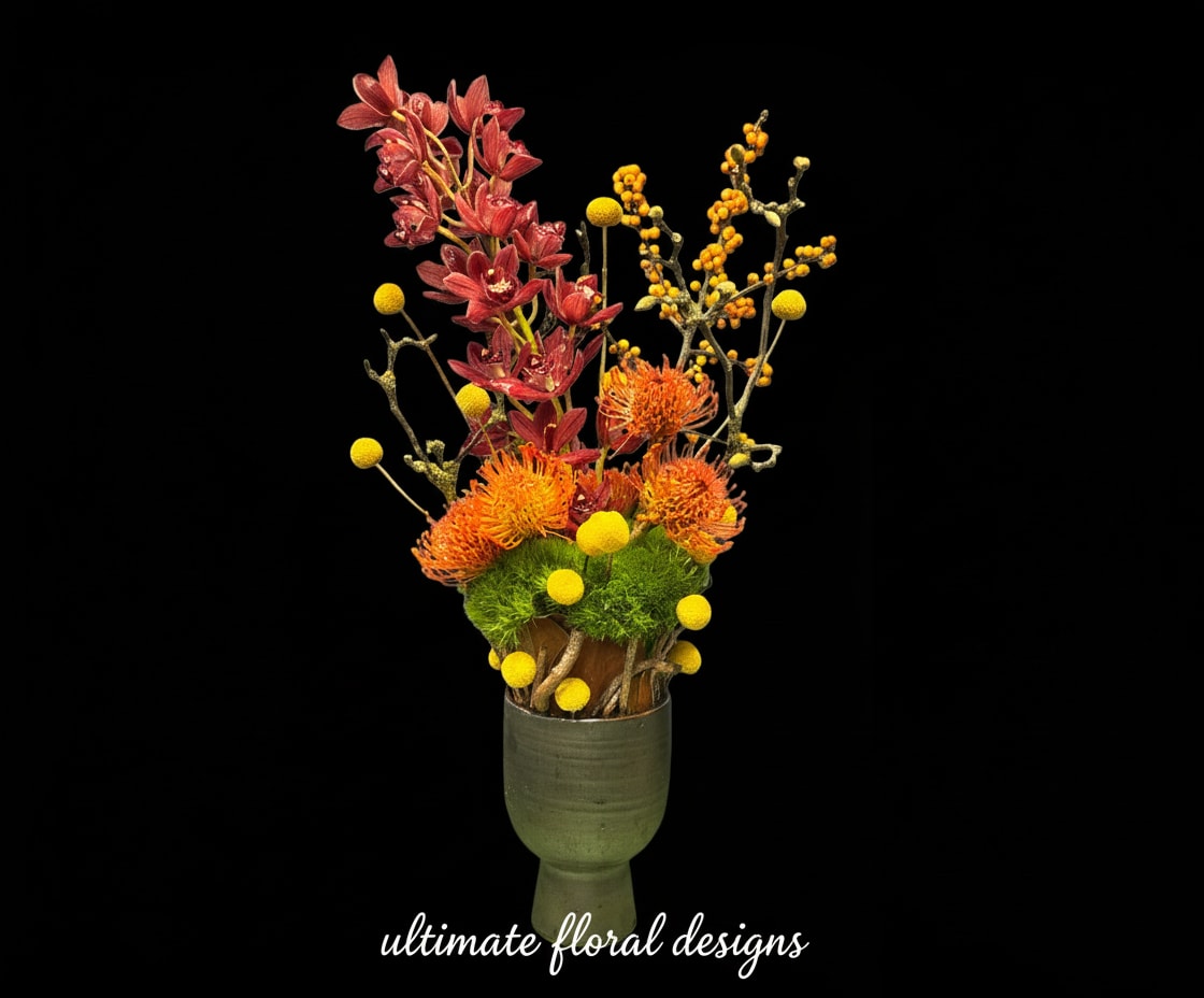 Golden Ember Orchid Arrangement - A striking modern floral composition featuring rich rust-toned cymbidium orchids, vibrant orange pincushion protea, golden craspedia, and sculptural textural botanicals, all grounded with lush green moss in a handcrafted ceramic vessel. This artistic arrangement blends warmth, movement, and architectural elegance, making it ideal for luxury interiors, statement consoles, executive offices, or refined gifting.  Designed with premium, long-lasting blooms and a sculptural silhouette, Golden Ember brings bold sophistication and contemporary floral artistry into any space.