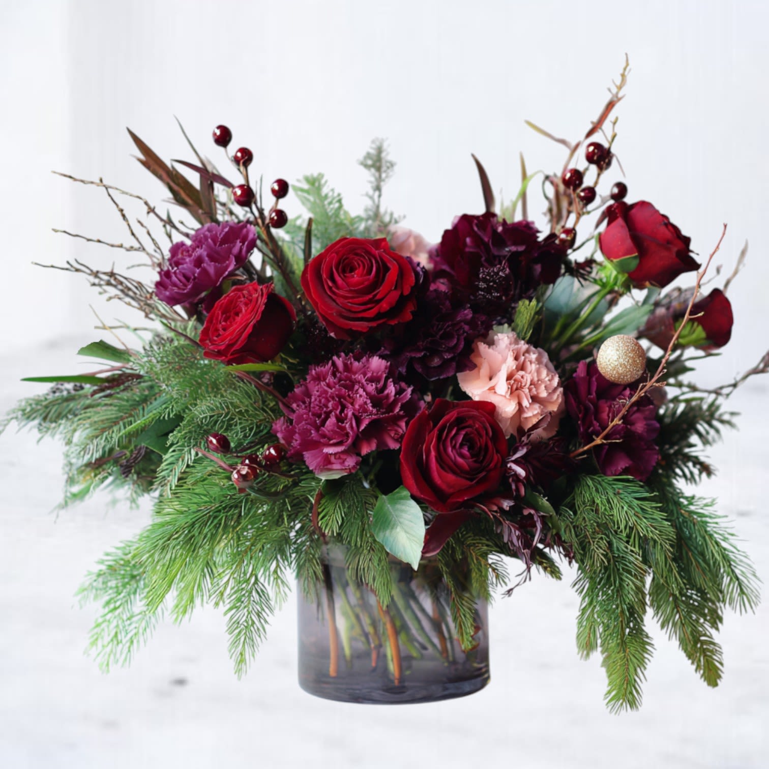 Crimson Evergreen - Rich, moody, and unmistakably winter, Crimson Evergreen is a jewel-toned holiday arrangement designed with depth, texture, and movement. This seasonal design features deep red roses, plum and wine-toned carnations, and soft blush accents, nestled into lush layers of fragrant evergreen boughs, winter berries, and natural branch elements.  Airy pine tips cascade gently over the vessel, giving the arrangement a relaxed, garden-style silhouette while maintaining a bold holiday presence. Subtle metallic accents add just enough seasonal shimmer without overpowering the organic feel. Designed in a smoky glass cylinder, Crimson Evergreen is perfect for those who love classic Christmas colors with a modern, editorial edge.