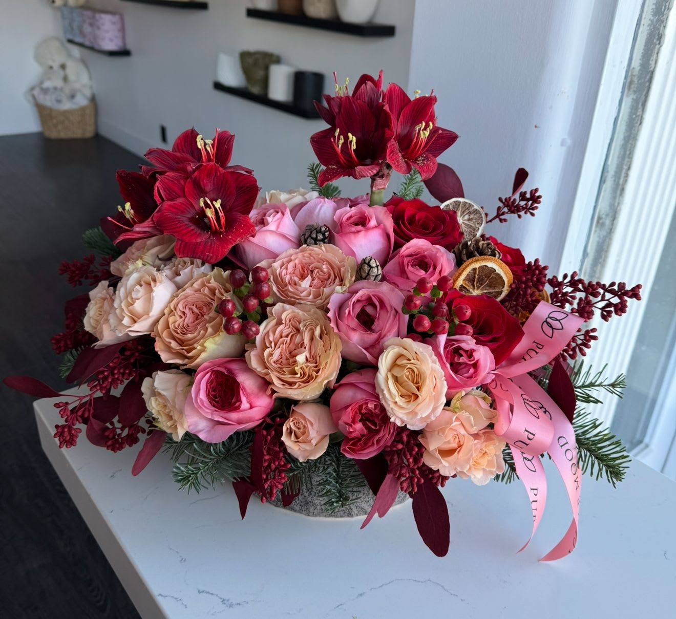 Holiday Amour Vase - Celebrate the season with the vibrant beauty of our Holiday Amour arrangement. Featuring bold red amaryllis, delicate garden roses, and soft pink accents, this masterpiece is complemented by festive greenery, berries, pinecones, and dried citrus for a touch of holiday charm. Finished with a luxurious pink ribbon, it’s the perfect centerpiece for your holiday celebrations or a heartfelt gift for loved ones.  	•	Premium blooms in red, pink, and peach tones 	•	Seasonal accents: berries, pinecones, dried citrus 	•	Elegant pink ribbon and festive greenery 	•	Handcrafted to add warmth and joy to any space 