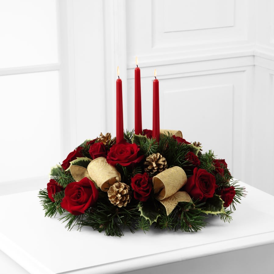 The  Celebration of the Season Centerpiece - The Celebration of the Season Centerpiece is a grand display of holiday elegance. Red roses and spray roses pop against a backdrop of assorted holiday greens and variegated holly that beautifully encircle three red taper candles. Accented with gold pine cones and gold metallic brocade ribbon this centerpiece creates a warm and enchanting glow to benefit their holiday festivities.