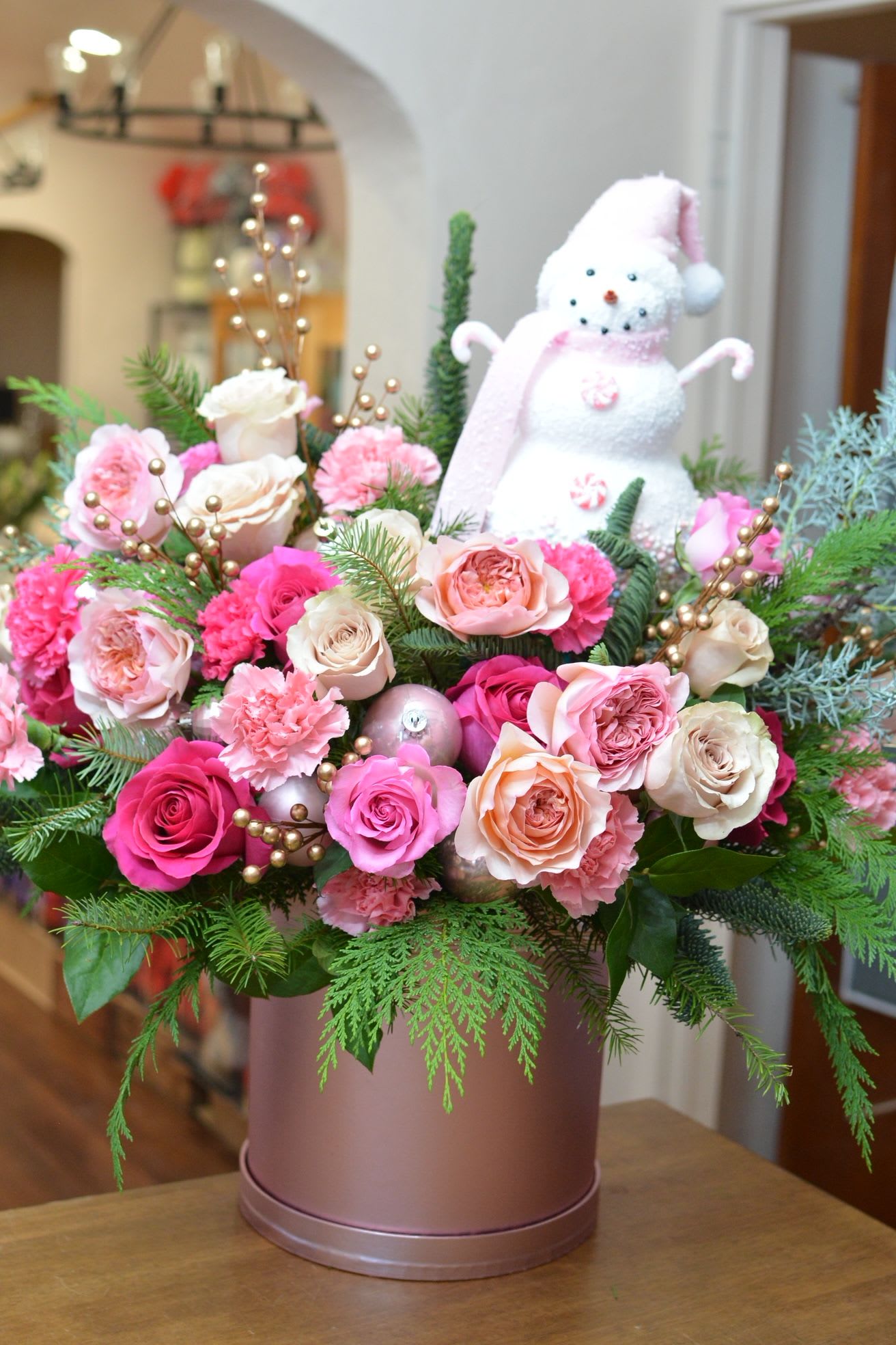 Blushing Snowfall Snowman - Make a bold holiday statement with Blushing Snowfall Snowman! This eye-catching Christmas arrangement showcases an adorable pink-and-white snowman surrounded by lush winter greenery, snowy white accents, vibrant hot pink highlights, and soft blush tones. Frosted textures, sparkling details, and playful pops of color create a festive look that’s both whimsical and glamorous. Perfect for brightening up your holiday décor, this arrangement brings cheerful charm, cozy winter magic, and a fun splash of pink to the Christmas season.