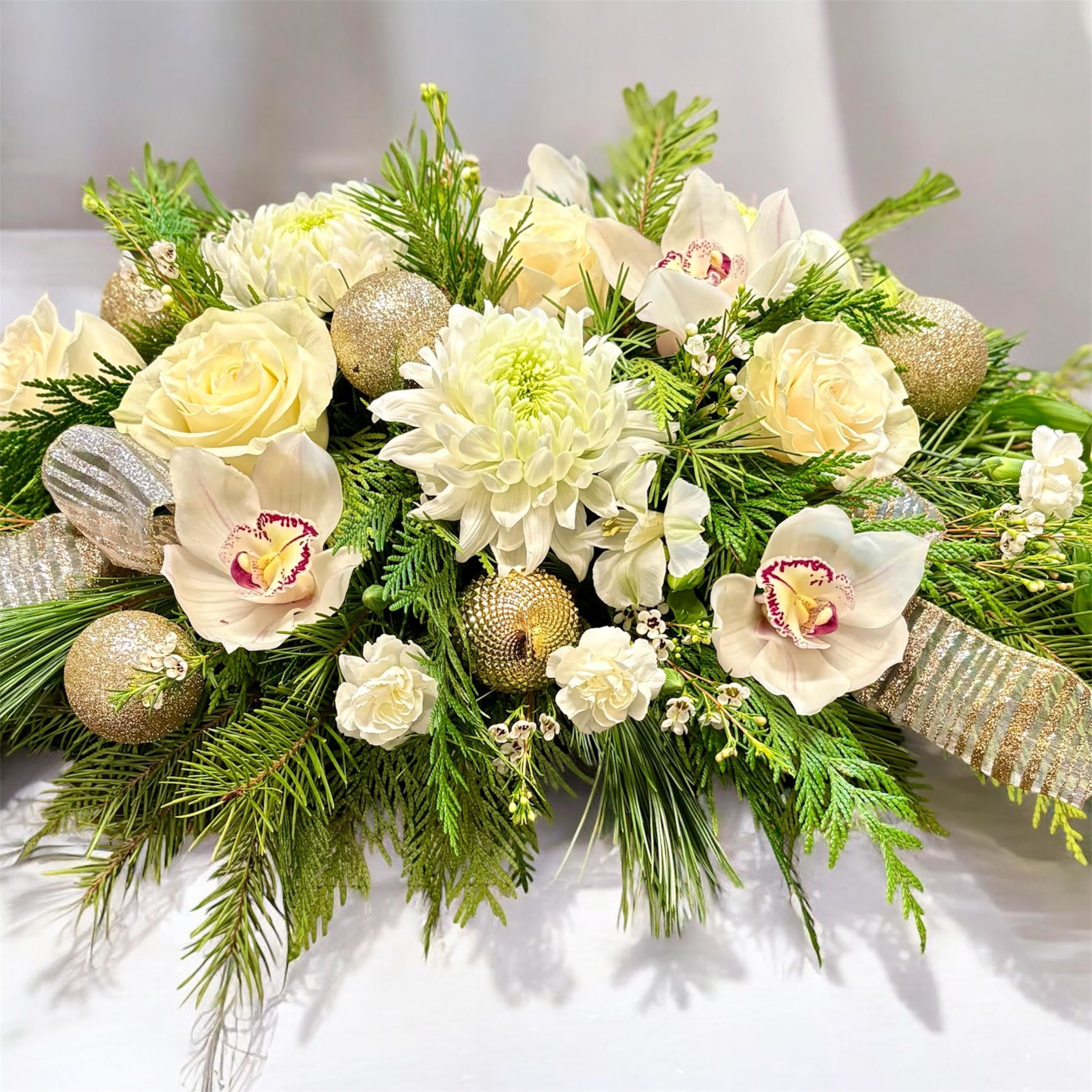 Ivory &amp; Gold Noel - A refined table centerpiece featuring soft cream and white flowers, artfully arranged to create a look of timeless elegance. Lush blooms are layered with delicate textures and gentle movement, while gold accents add a warm, festive glow. Designed low and balanced, this centerpiece enhances the table without overwhelming it, making it perfect for holiday gatherings or special celebrations. The overall feel is luxurious yet inviting, blending classic winter beauty with subtle seasonal sparkle.