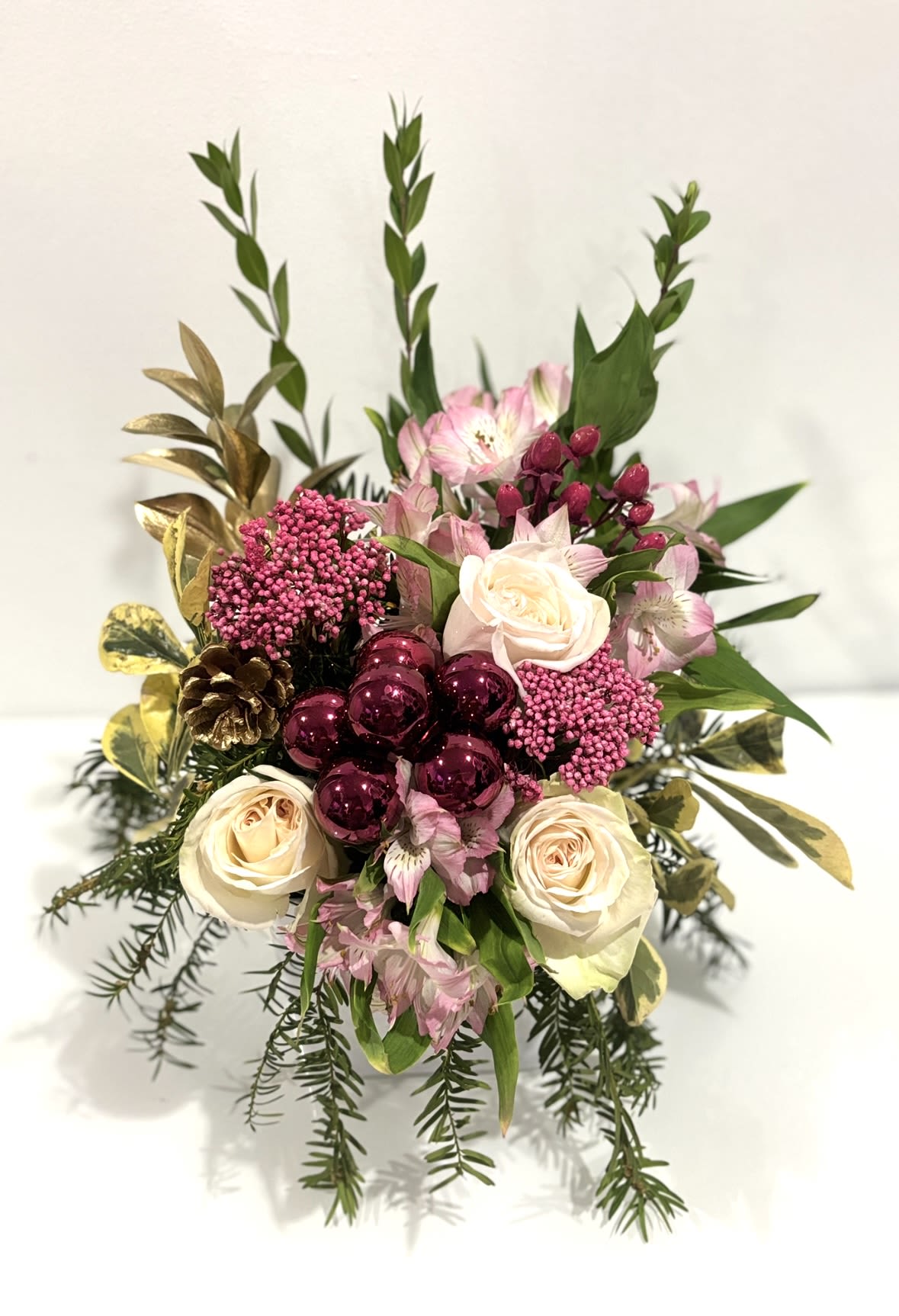 Blush Winter Elegance - A refined festive floral arrangement featuring soft pink and cream roses, pink alstroemeria, and delicate clusters of blush blooms, accented with fresh pine greenery. Shimmering silver ornaments and a subtle gold pinecone add a graceful holiday touch, all beautifully presented in a classic white vessel-perfect for Christmas and winter celebrations. 
