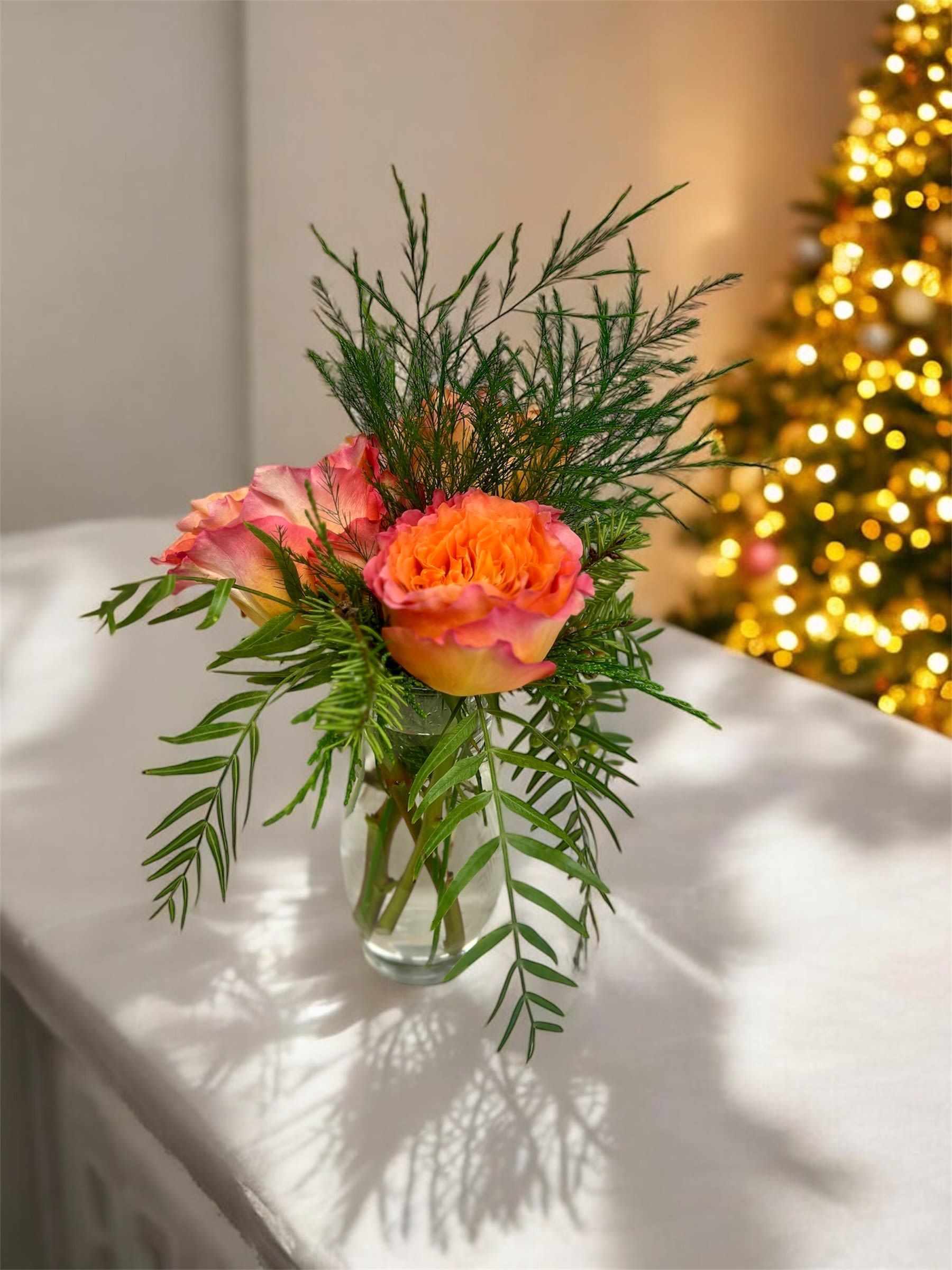 Sunset Whisper - Sunset Whisper is a delicate, modern arrangement designed to capture the warmth and quiet beauty of golden hour. Featuring premium garden roses in soft peach and coral tones, this petite design is accented with airy seasonal greenery and thoughtfully arranged in a clear glass vase for a clean, organic finish.  Perfect for everyday gifting, intimate celebrations, or simply brightening a space, Sunset Whisper offers understated elegance with a gentle, romantic presence.  