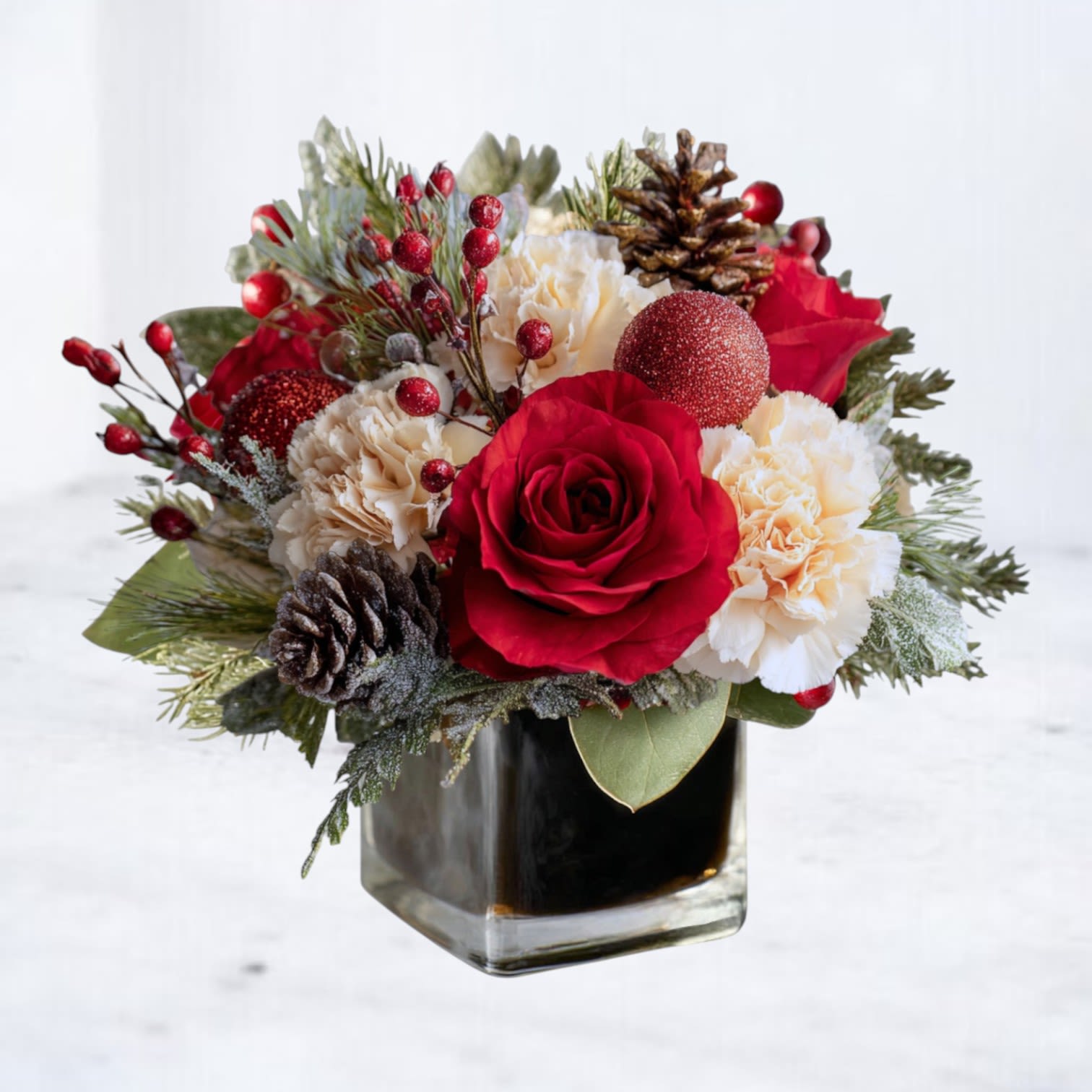 Merry Munchkin — Holiday Favorite - A Festive Mini Arrangement for Big Holiday Cheer  Bring a burst of joyful holiday spirit into any room with the Merry Munchkin — a small but mighty Christmas arrangement designed to brighten desks, countertops, nightstands, mantels, and holiday tables. This cheerful mini bouquet blends velvety red roses, fluffy white carnations, and seasonal winter greenery, all arranged in a black lined, 4x4 cylinder vase for a modern, high-contrast holiday look.  Designed to deliver maximum merry in a compact size, this is the perfect gift for teachers, coworkers, neighbors, dinner hosts, or anyone who deserves a festive surprise. Small enough to fit anywhere. Cute enough to steal the show.