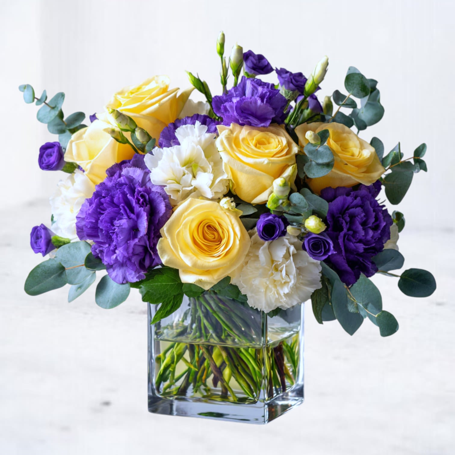 Luminous Contrast - Luminous Contrast is a vibrant, modern arrangement designed to uplift and energize any space. Featuring a striking blend of golden yellow roses, rich purple carnations, and soft white accents, this design is balanced with fresh eucalyptus and seasonal greenery for a clean, contemporary finish.  Arranged in a clear glass cube that showcases natural stem movement, Luminous Contrast offers a bold yet approachable look — perfect for birthdays, congratulations, encouragement, thank-you gifts, or simply brightening someone’s day.  This arrangement is ideal for recipients who appreciate color-forward design with a refined, polished feel.