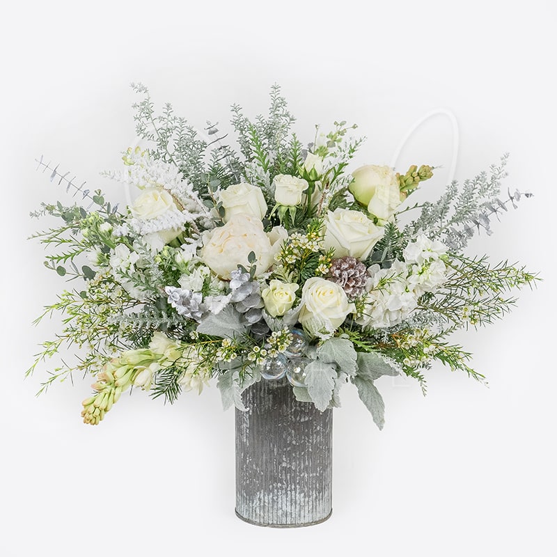 Silver Bells - A holiday favorite that’s ready to wow! Experience the magic of the season with this stunning all-white &amp; silvery grey floral arrangement adorned with seasonal greenery and festive accents.  Featured in our Metal Norah Vase 7.5" x 4.5" (ONE SIDED)
