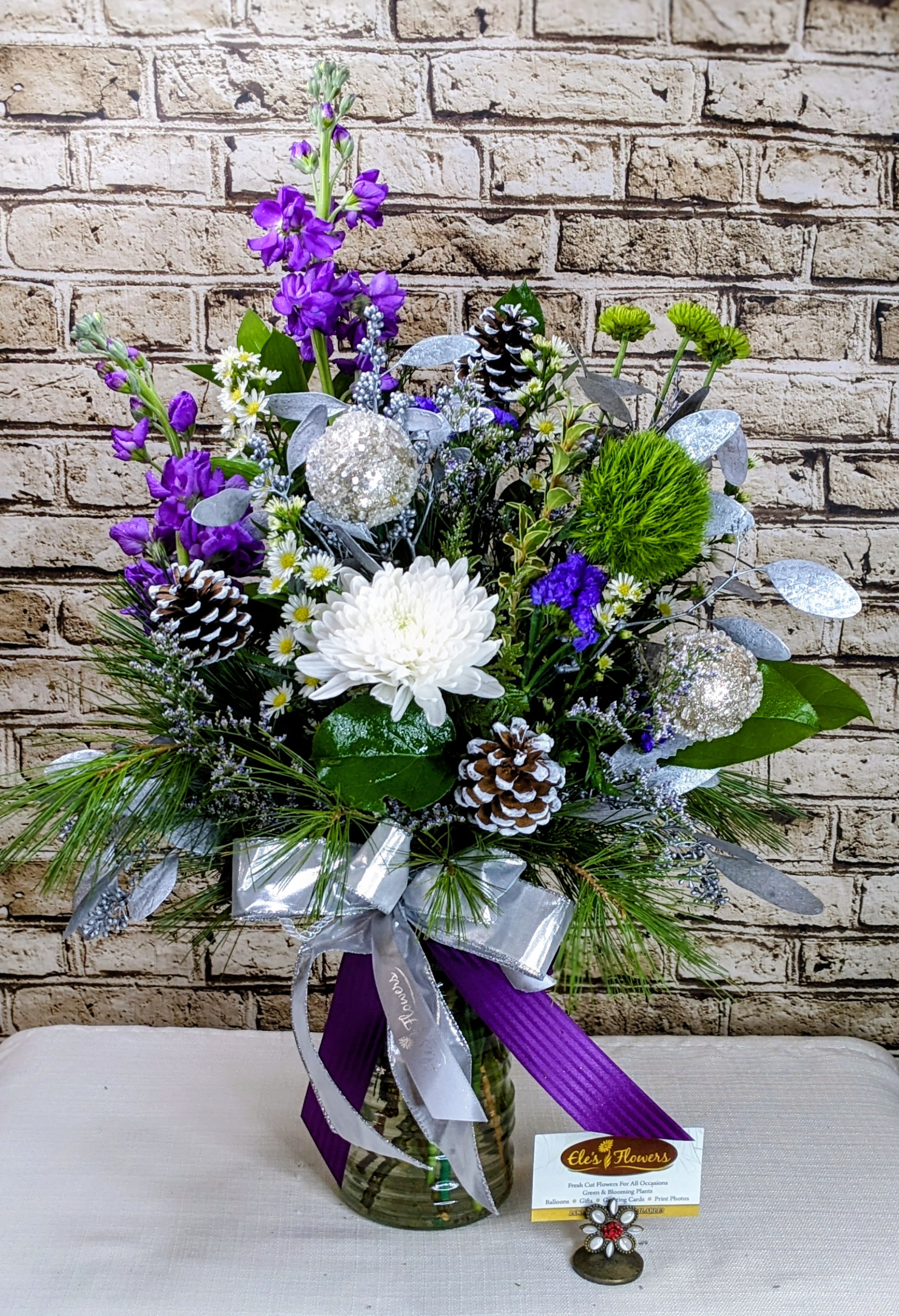 Amethyst Winter  - Step into a winter wonderland with Amethyst Winter, a show-stopping floral arrangement that blends rich jewel tones with sparkling seasonal accents. Lush purple blooms rise gracefully above crisp white florals, creating a striking contrast that feels both elegant and enchanting. Frosted pinecones, shimmering silver ornaments, and cool metallic foliage add depth and texture, while fresh evergreen touches bring in that unmistakable holiday freshness.  Finished with a flowing silver and deep-purple ribbon, this design feels luxurious, festive, and effortlessly sophisticated. It’s the kind of arrangement that stops people in their tracks—perfect for holiday entertaining, winter celebrations, or gifting someone who deserves something truly special.  Blooms and embellishments may vary.   Approximate Dimensions: 26"H x 18"W