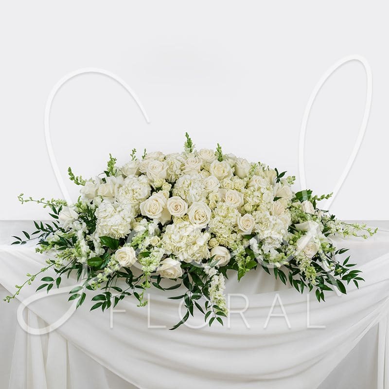 Funeral Casket Spray - A neutral floral spray design with a mix of premium and seasonal white blooms accented by mixed greenery and textural elements. Designed in a natural and classic style on a saddle base to place atop a casket.