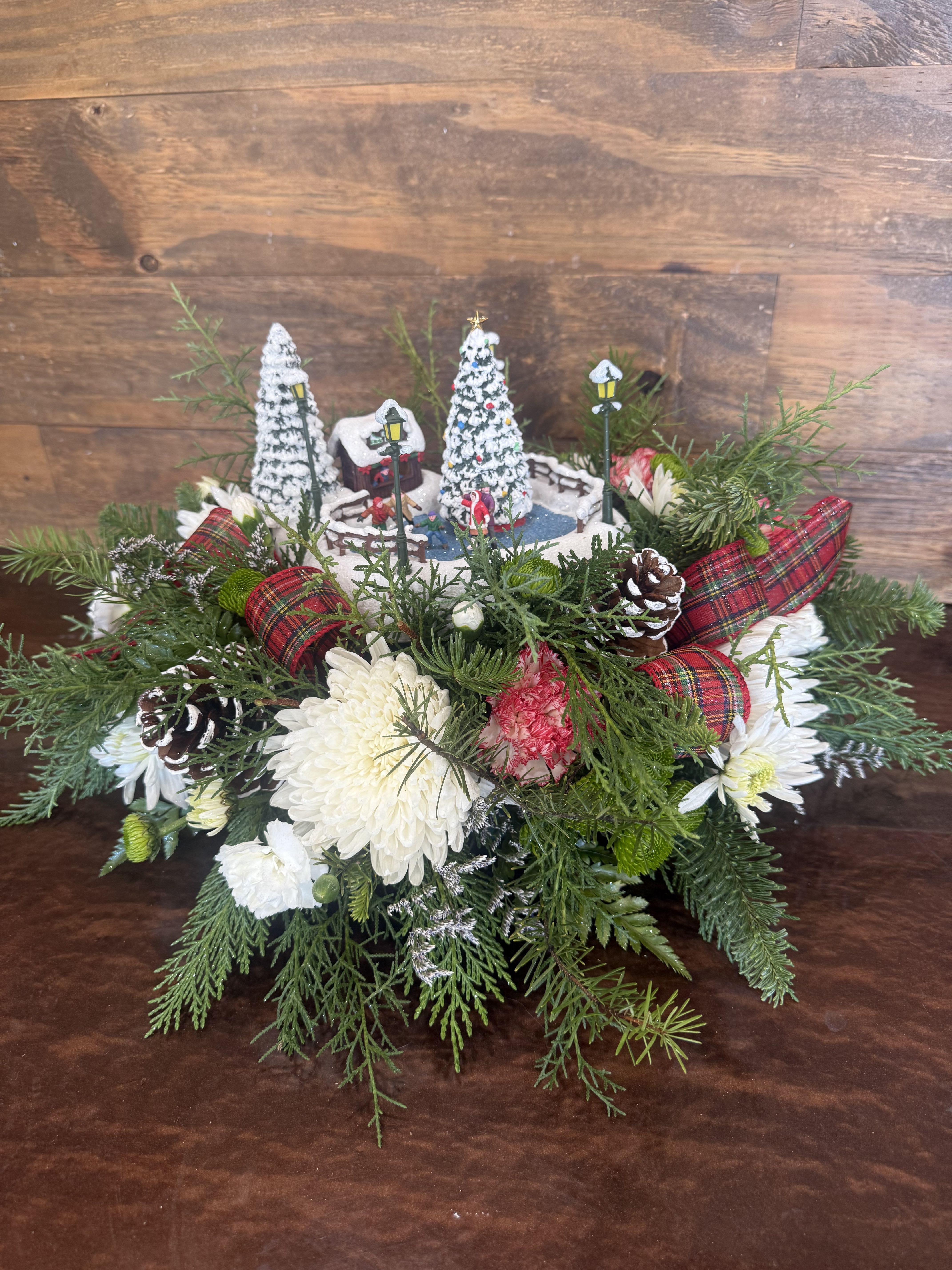 Frosty Village Centerpiece - This enchanting Christmas centerpiece captures the magic of a winter village scene nestled in lush, fresh florals. At its heart is a whimsical miniature ice-skating pond, complete with tiny skaters gliding gracefully, surrounded by snow-flocked evergreen trees adorned with colorful lights and topped with glowing stars. Vintage-style street lamps cast a soft, warm glow over the frosty landscape, evoking nostalgic holiday charm.