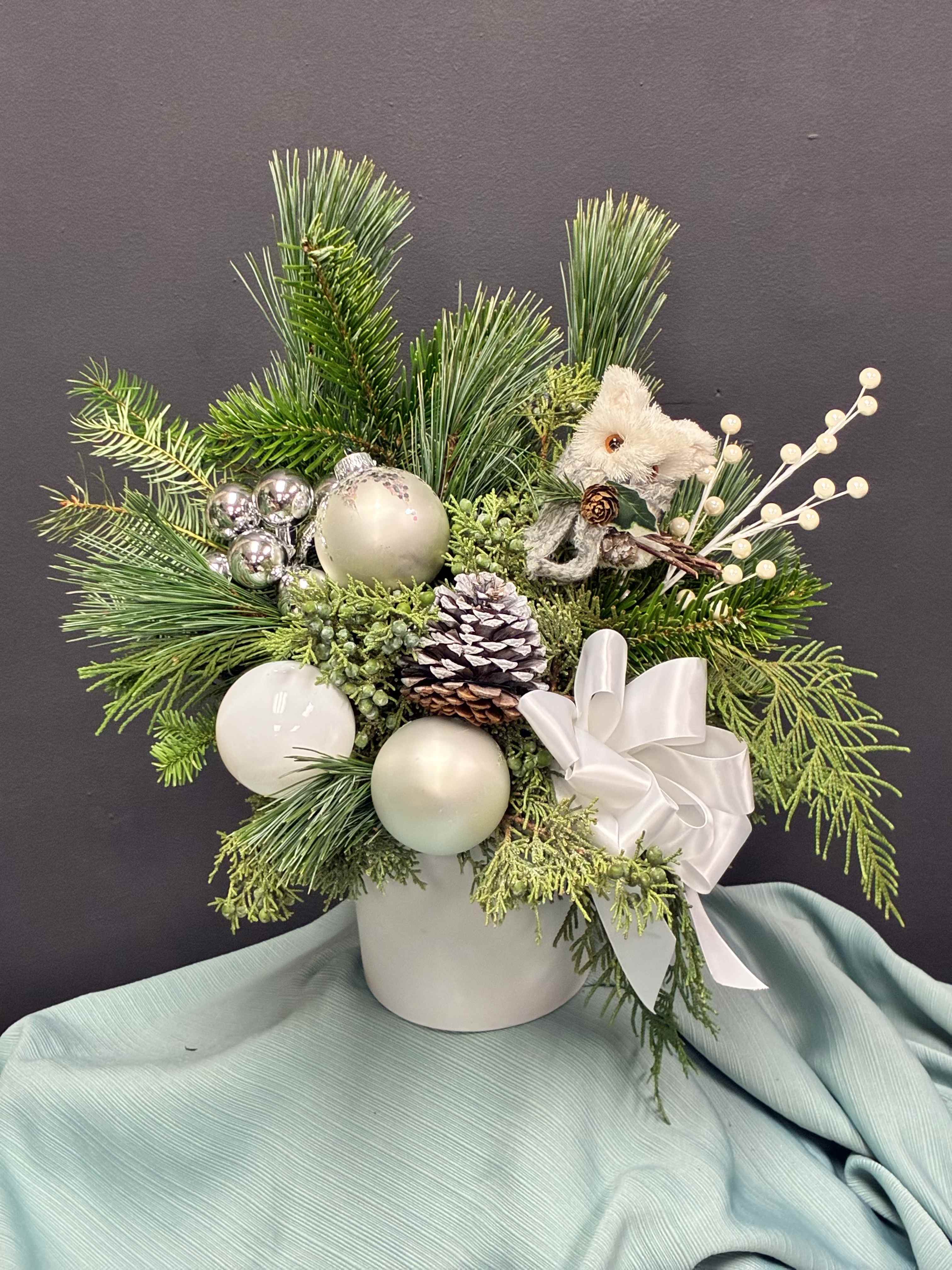 Winter Snuggle - This heartwarming holiday arrangement blends winter elegance with a touch of whimsy. Set in a white ceramic container, it features lush pine and fir branches accented with frosted pinecones, white berry sprays, and shimmering silver baubles. A soft white ribbon bow adds a graceful flourish, while the centerpiece—a plush white bear—brings a cozy, nostalgic feel. The cool tones of green, white, and silver evoke a peaceful winter morning, making this piece perfect for holiday décor or a comforting seasonal gift.  **holiday decor may vary** you can let us know your color preference and we will do our best to accommodate 