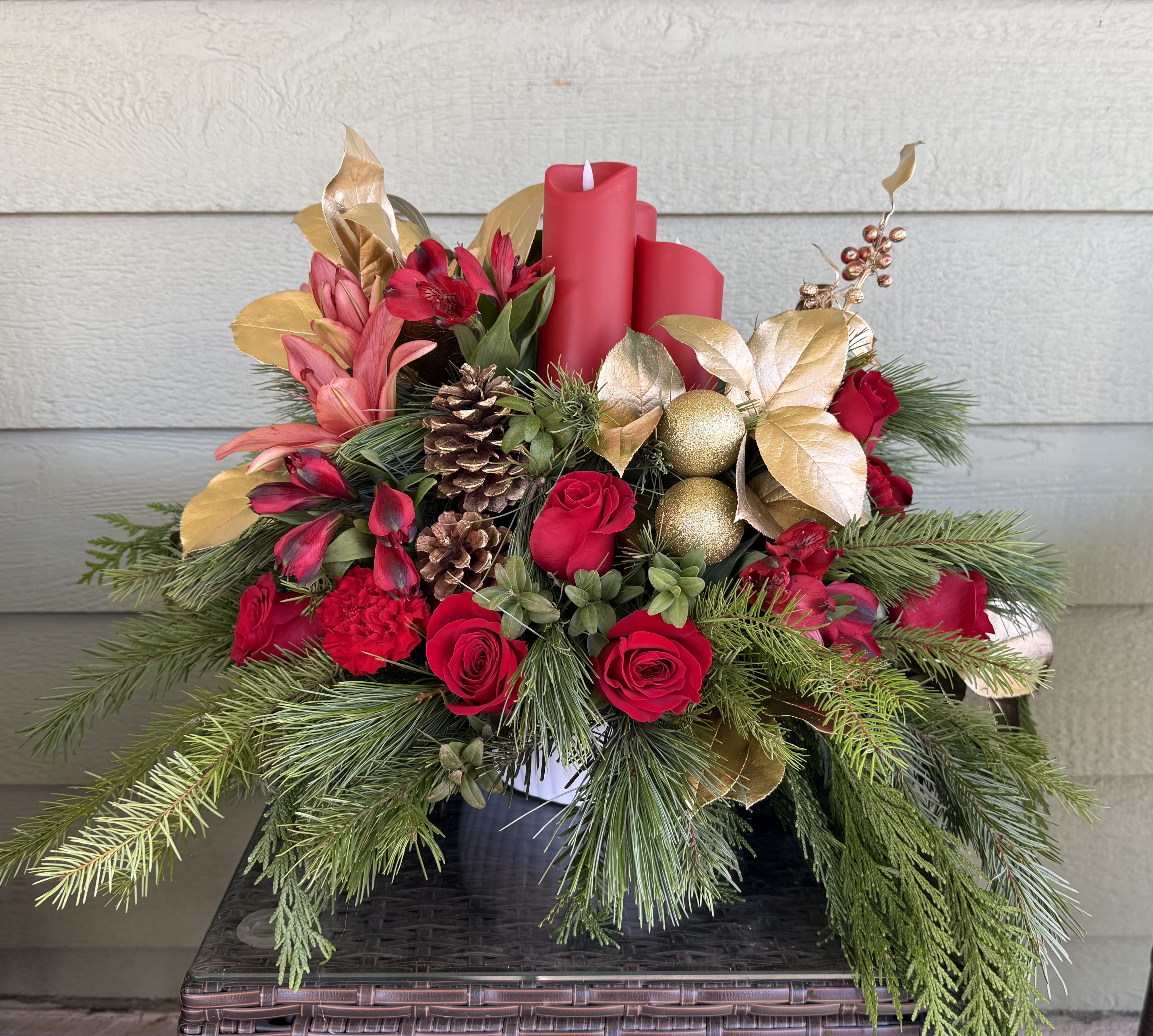 Elegant Holiday Evergreen Centerpiece - Celebrate the season with this lush holiday centerpiece, artfully designed with fresh evergreen foliage, velvety red roses, and festive accents. Three red pillar candles rise from a bed of fragrant pine, cedar, and fir, surrounded by natural pinecones, gold ornaments, and warm metallic foliage for a classic yet elevated look. Cascading greenery adds graceful movement, making this arrangement a stunning focal point for dining tables, entryways, or mantel displays.  Perfect for holiday gatherings, seasonal décor, or gifting, this centerpiece brings timeless Christmas charm and cozy sophistication to any space.  Approximate size: Low and full with flowing greenery Care: Keep greenery lightly misted and away from direct heat