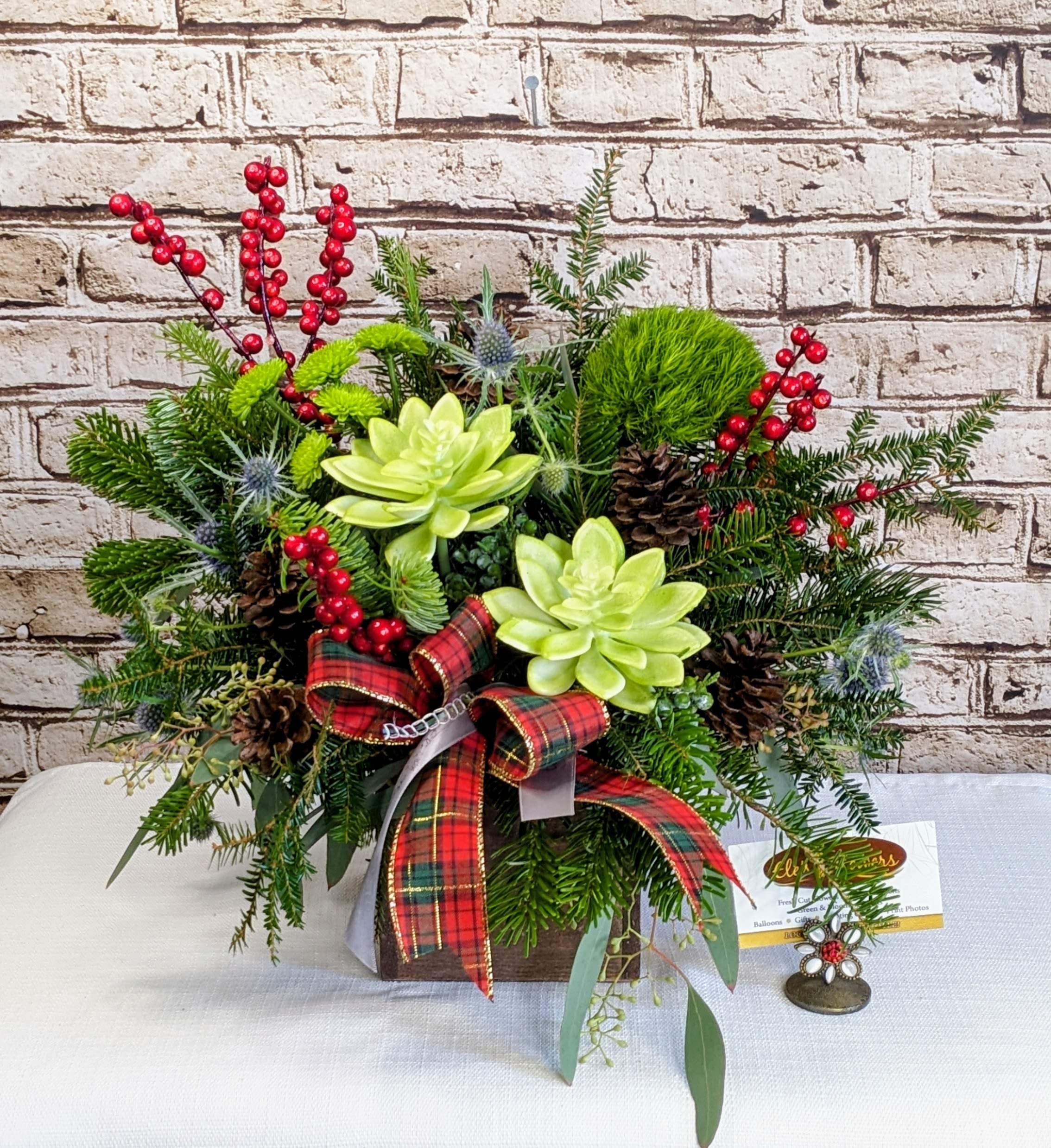 Winter Garden - This festive arrangement is a cheerful celebration of winter charm and holiday spirit. Lush evergreen textures create a full, cozy foundation, accented with pops of bright red for a classic seasonal feel. Playful touches of natural elements add warmth and woodland whimsy, while soft green focal accents bring a fresh, modern twist. Finished with a bold plaid ribbon for extra holiday flair, this piece feels both rustic and joyful—perfect for spreading Christmas cheer on a table, counter, or as a thoughtful seasonal gift.  Approximate Dimensions: 15"H x 14"W