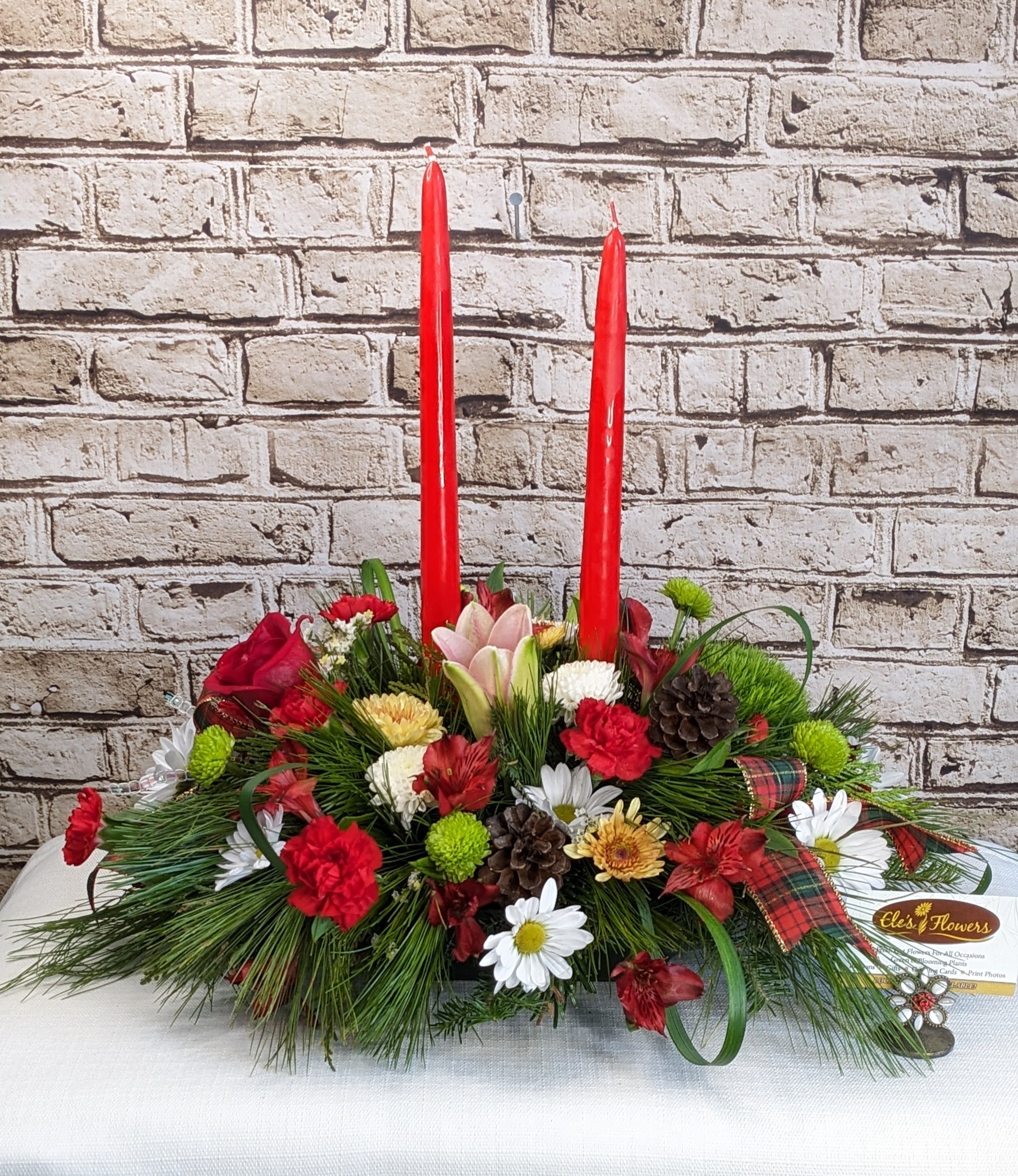 Merry &amp; Bright Holiday Centerpiece - True to its name, Merry &amp; Bright is a joyful holiday centerpiece that brings instant cheer to any table. Vibrant reds, crisp whites, and fresh pops of green come together in a lush seasonal design that feels classic, cozy, and full of Christmas spirit. Warm natural textures add a charming woodland touch, while festive accents and glowing candles create a welcoming holiday glow. Finished with a playful plaid ribbon, this centerpiece is perfect for gatherings, gift-giving, or making your home feel extra merry all season long.  Approximate Dimensions: 21"L x 14"H